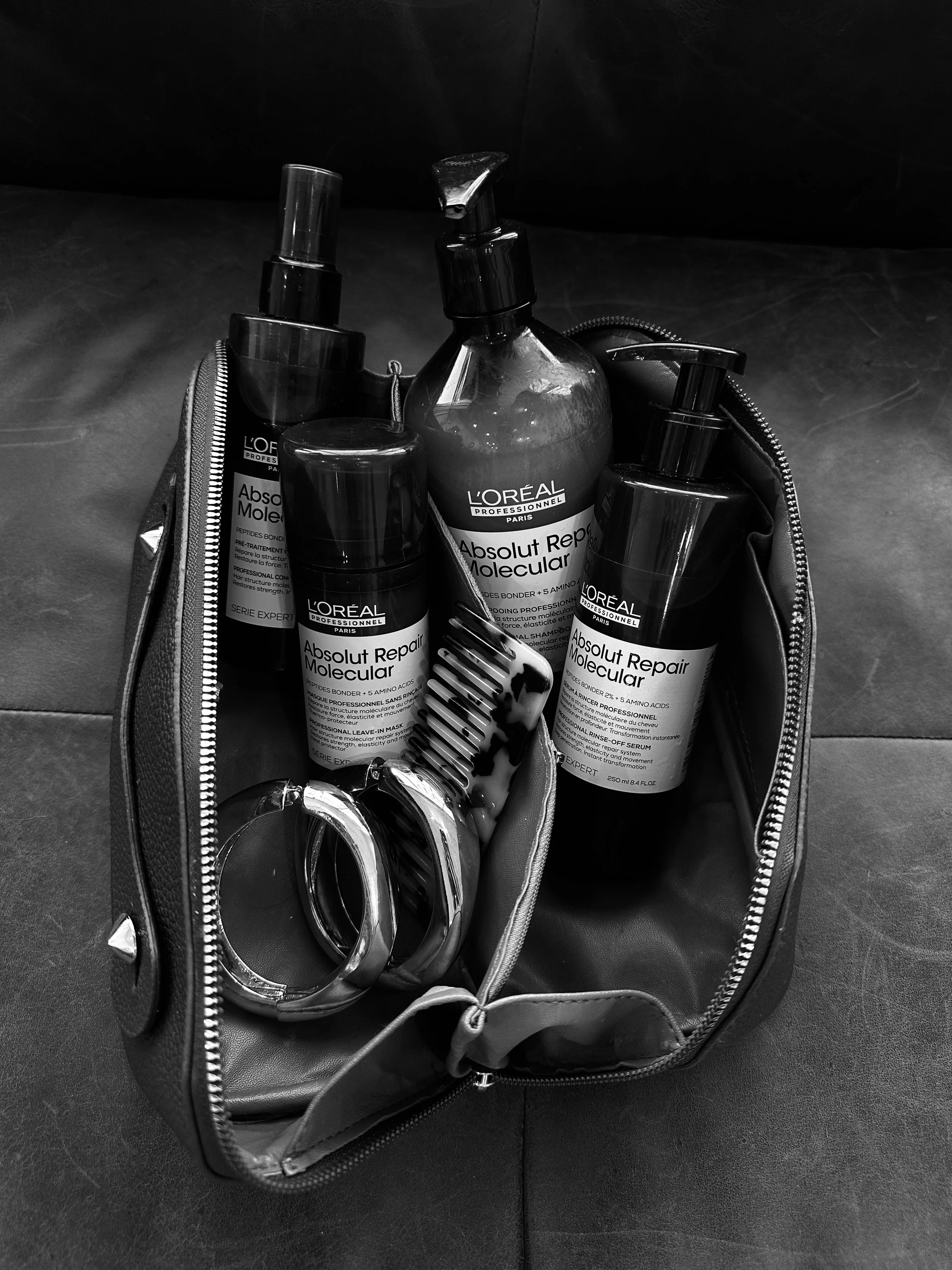 A black and white photograph of a small zipped bag containing hair care products, including bottles of L'Oreal Absolut Repair Molecular, and hair scissors.