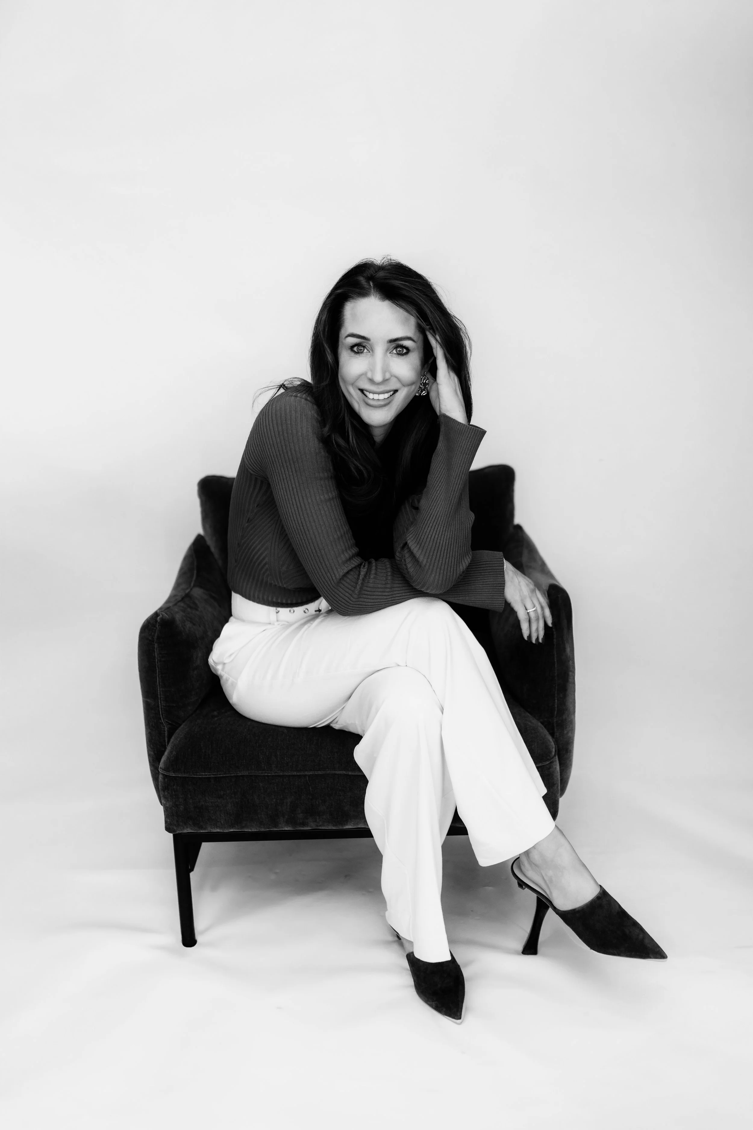 A woman sitting on a dark armchair, smiling at the camera, wearing a long-sleeve top, white pants, and high heels, with her hand on her head, in a studio with plain light background.