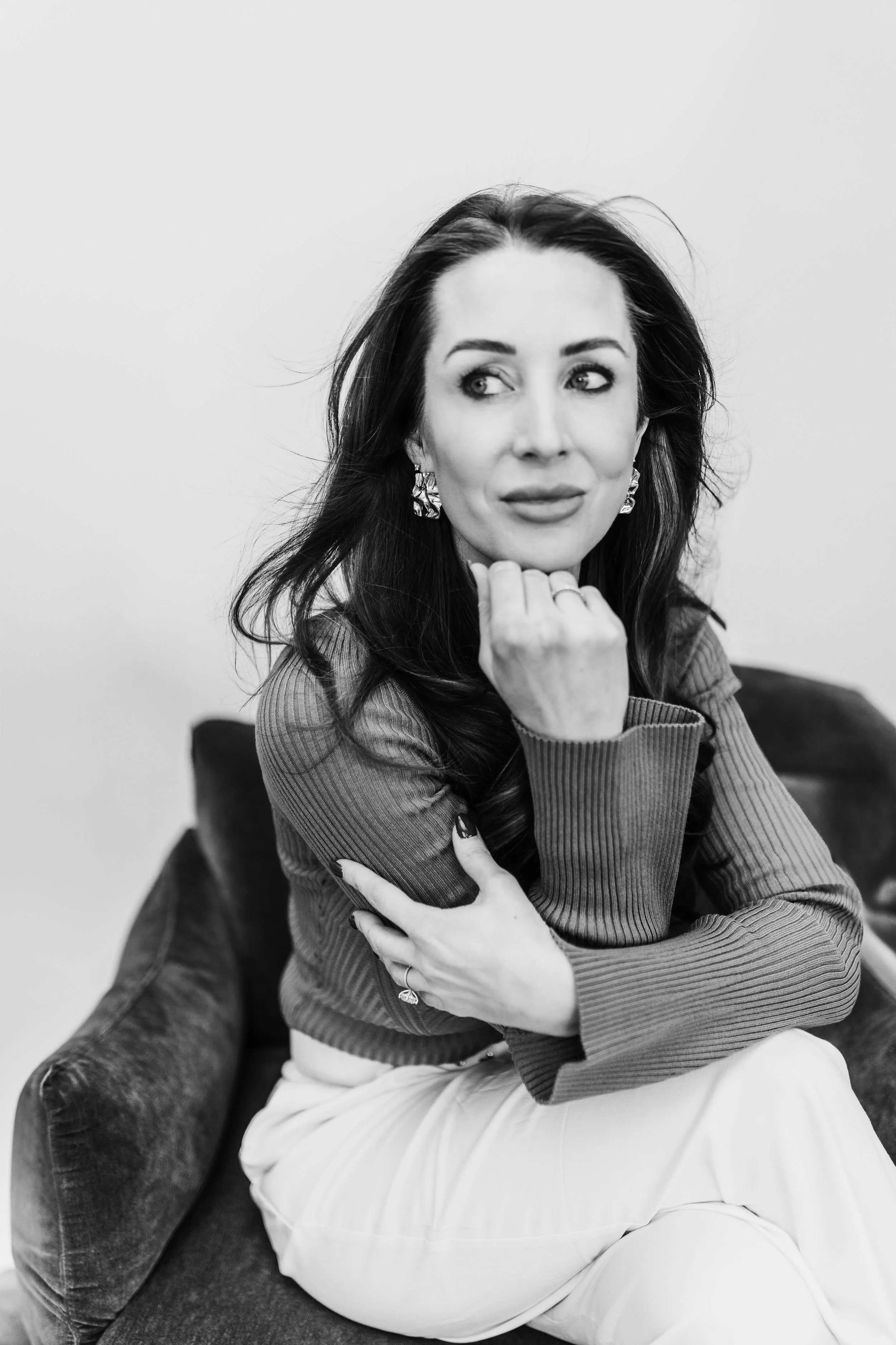 A black and white photo of a woman sitting on a dark-colored couch, with long dark hair, wearing a ribbed long-sleeve top and light-colored pants, looking off to the side with a thoughtful expression and her chin resting on her hand.