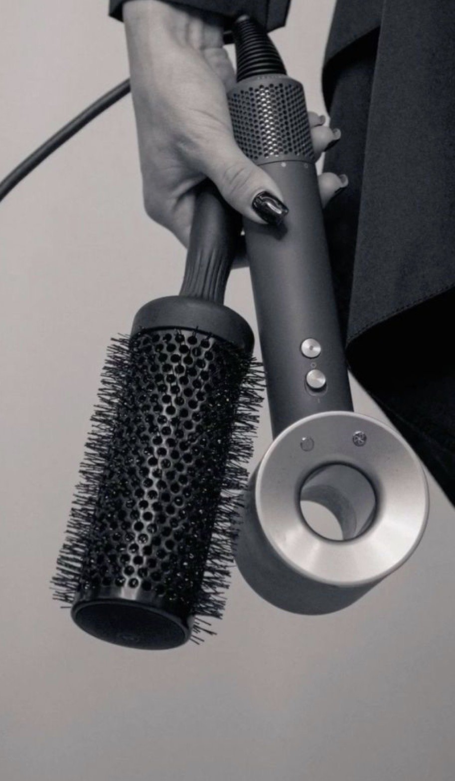 Close-up of a person holding a black round hairbrush and a gray cordless hair dryer with silver buttons and nozzle, against a plain background.