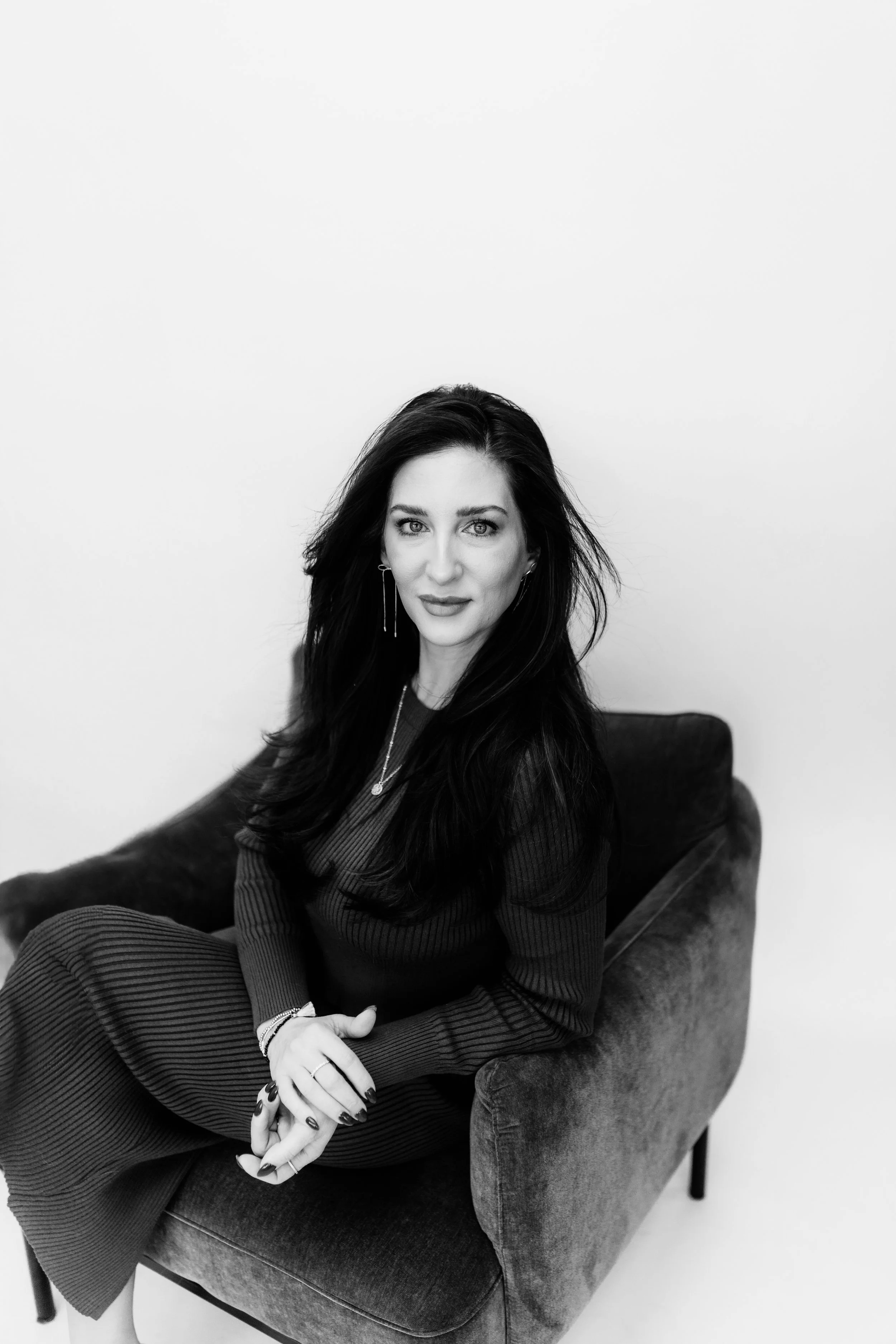 A woman with long dark hair sitting on a velvet armchair against a plain white wall, looking at the camera with a slight smile, holding her hands in her lap.