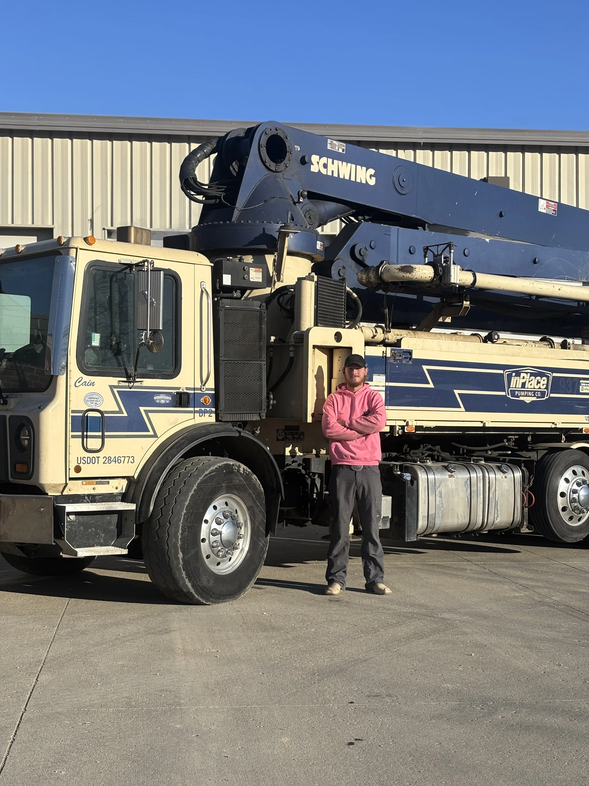 A man in a pink hoodie and dark pants standing with arms crossed in front of a large construction or utility truck with a crane arm marked 'Schwing'.