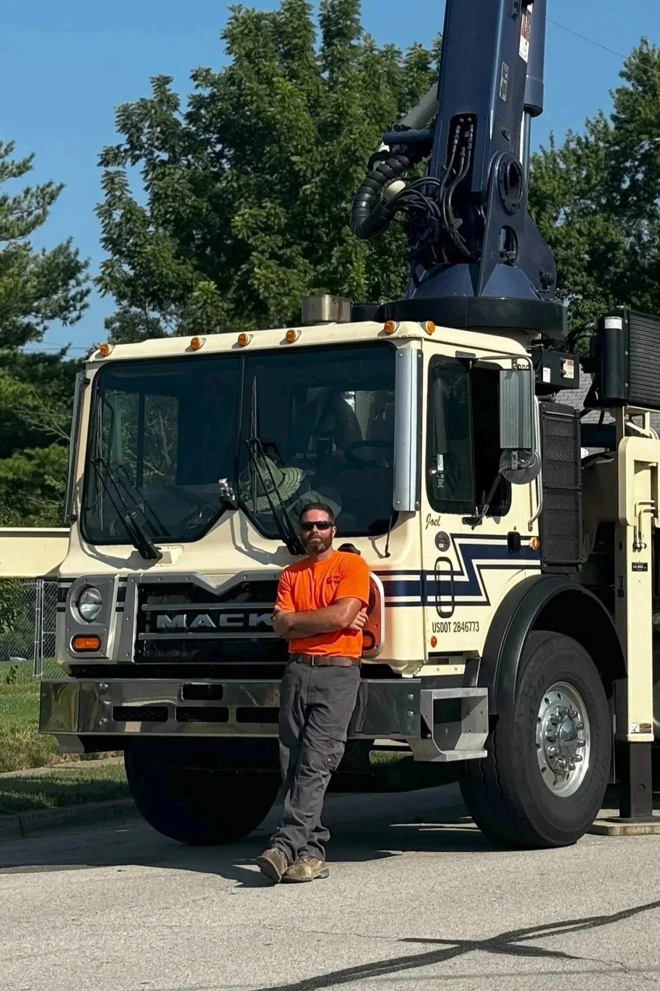A man wearing an orange shirt and gray pants standing with arms crossed in front of a large Mack truck with a crane attachment.
