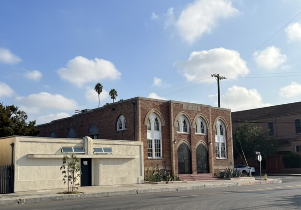 A brick building with arched windows and a small set of stairs leading to the entrance, alongside a white structure with a door and windows, set against a bright blue sky with scattered clouds and tall palm trees in the background.