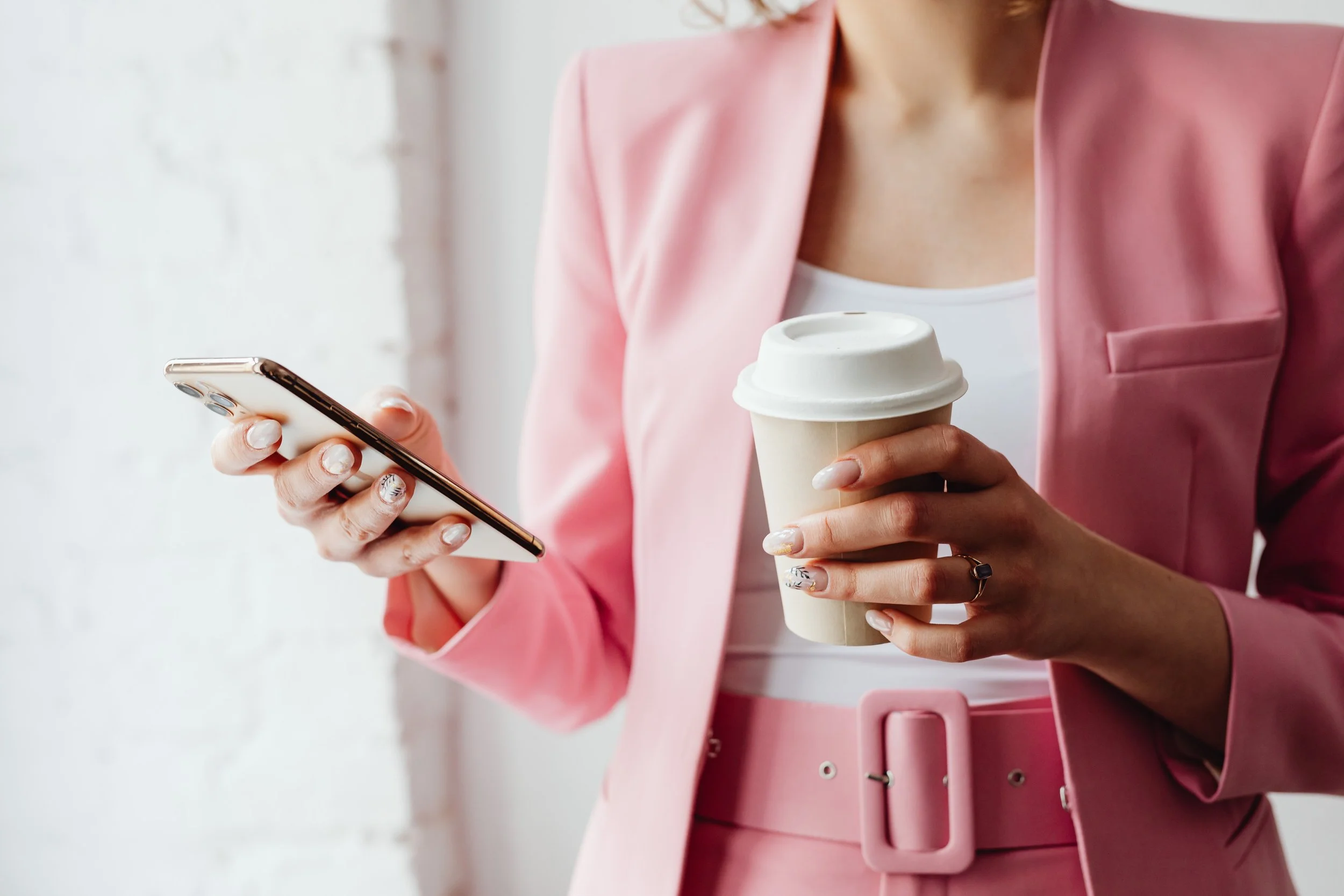 A woman in a pink blazer holding a coffee cup in one hand and a smartphone in the other, with a white wall in the background.