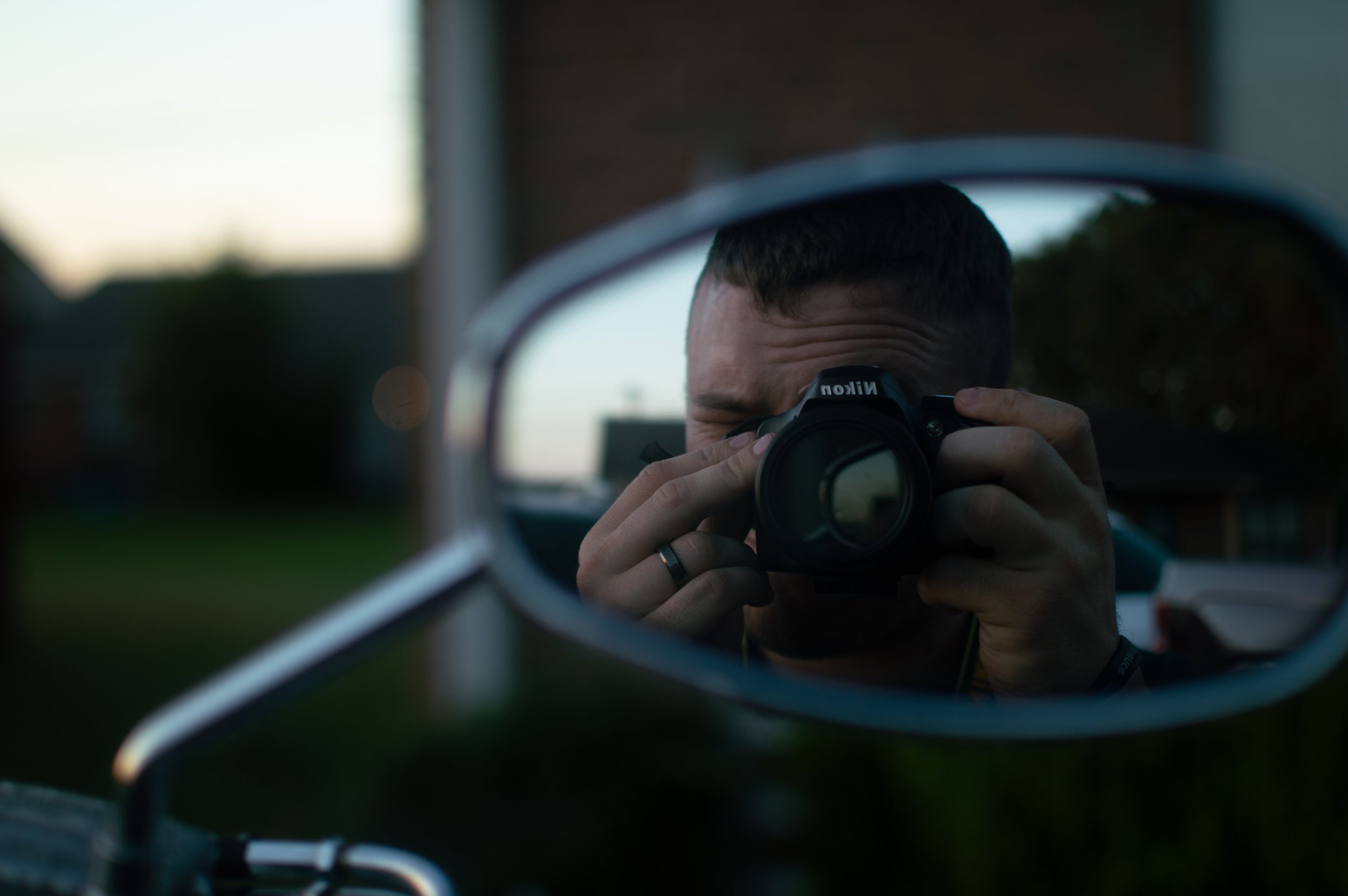 Person taking a photo of themselves in a car side mirror with a Nikon camera during sunset or twilight.