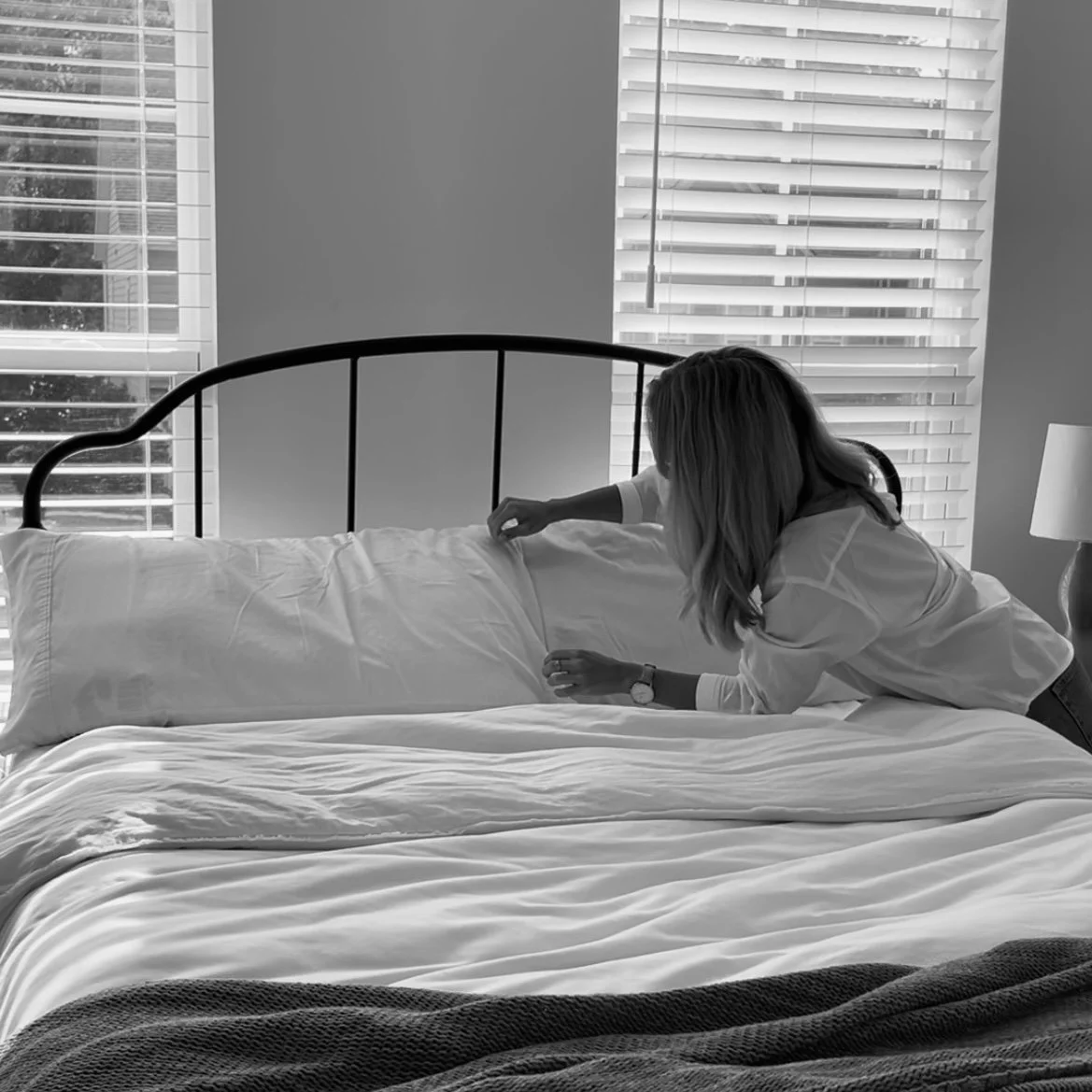 A woman arranging a pillow on a bed in a bedroom with closed blinds on windows, black-and-white photo.