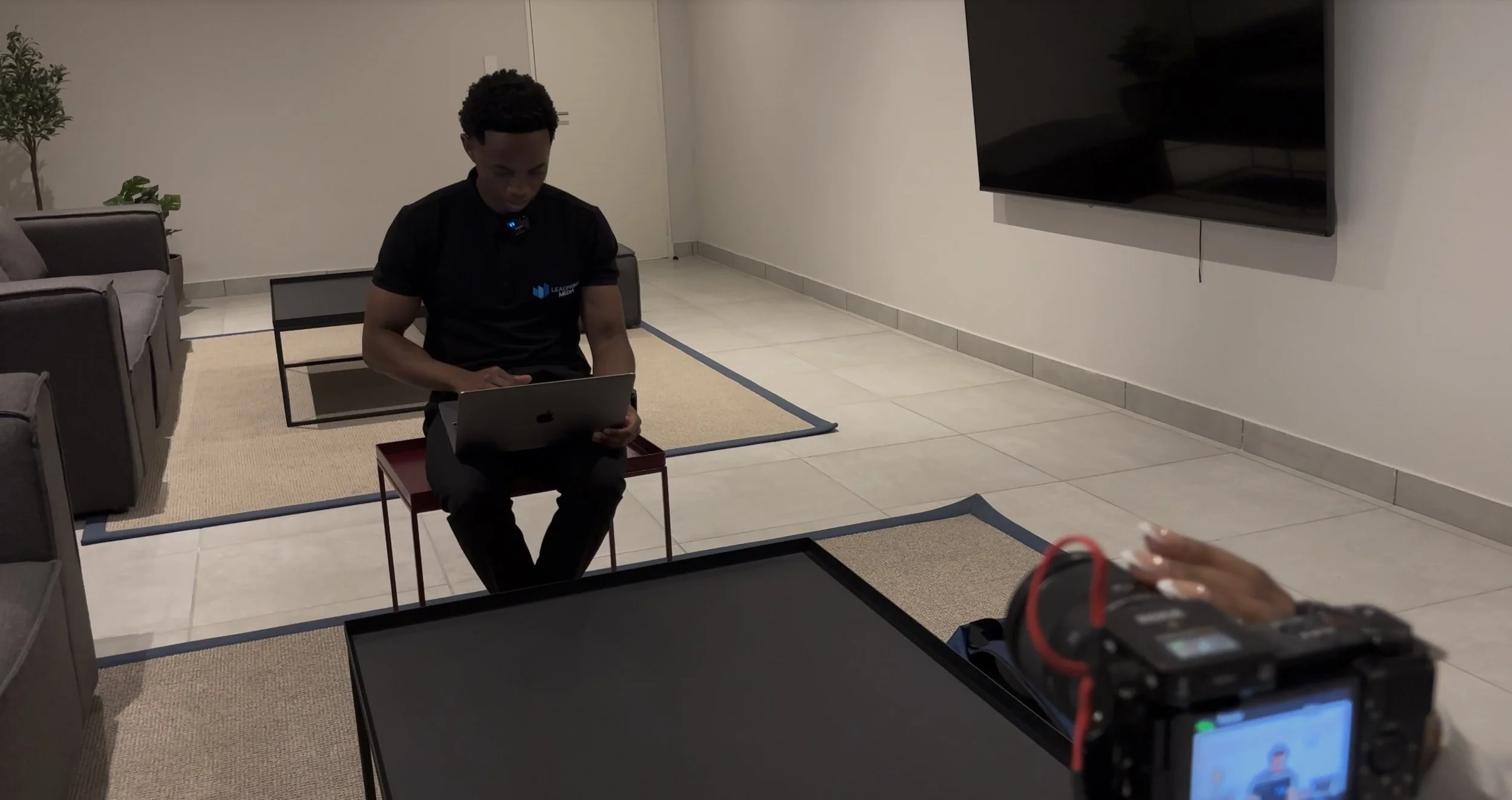 A person sitting on a red chair working on a laptop, with filming equipment in front, in a modern living room with white walls, a mounted flat-screen TV, a sofa, a coffee table, and a potted plant.