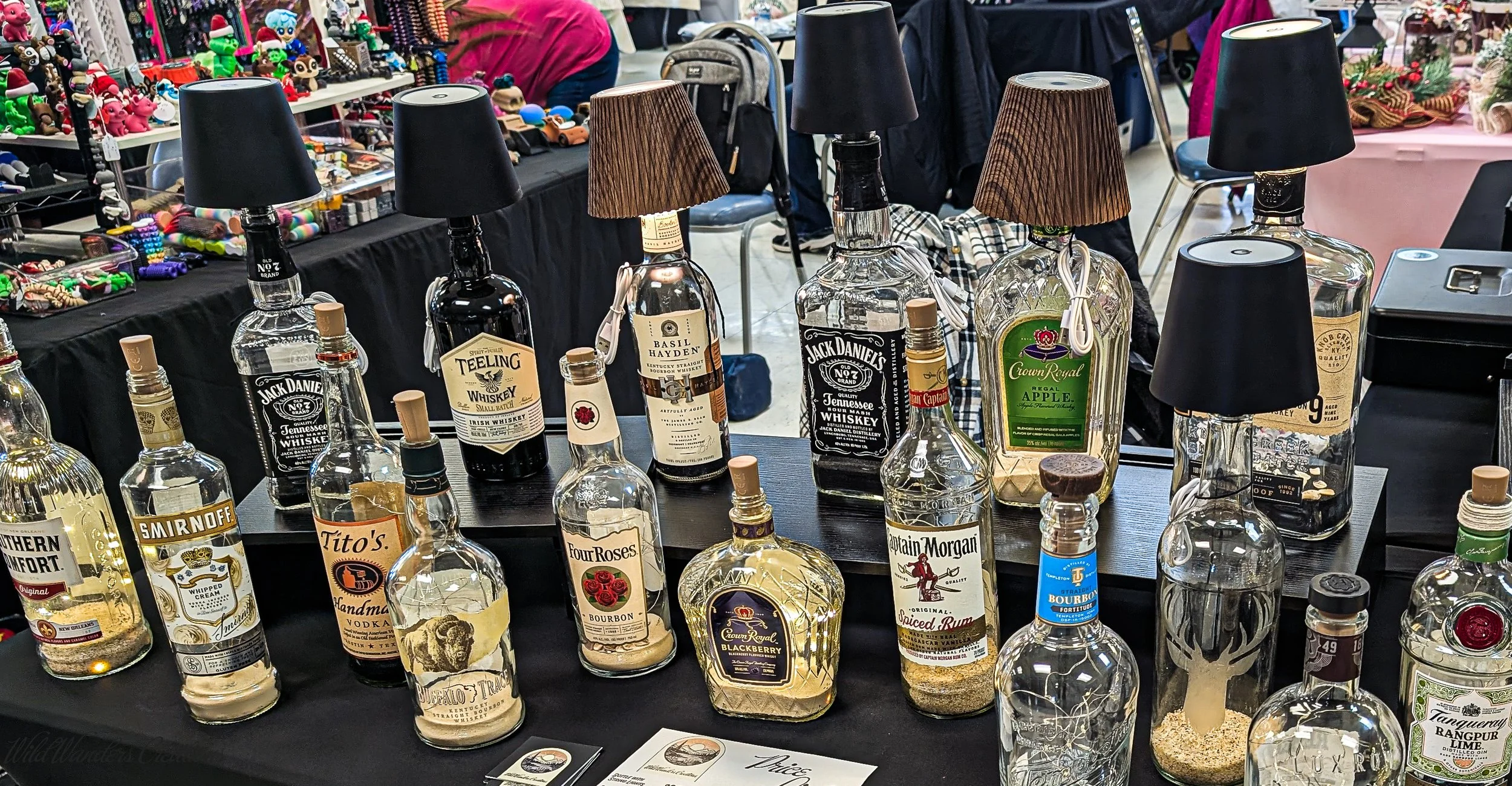 Table displaying various liquor bottles at a market or trade show, with black and brown shade lamps attached to some bottles, and a background with other vendor tables and shoppers.