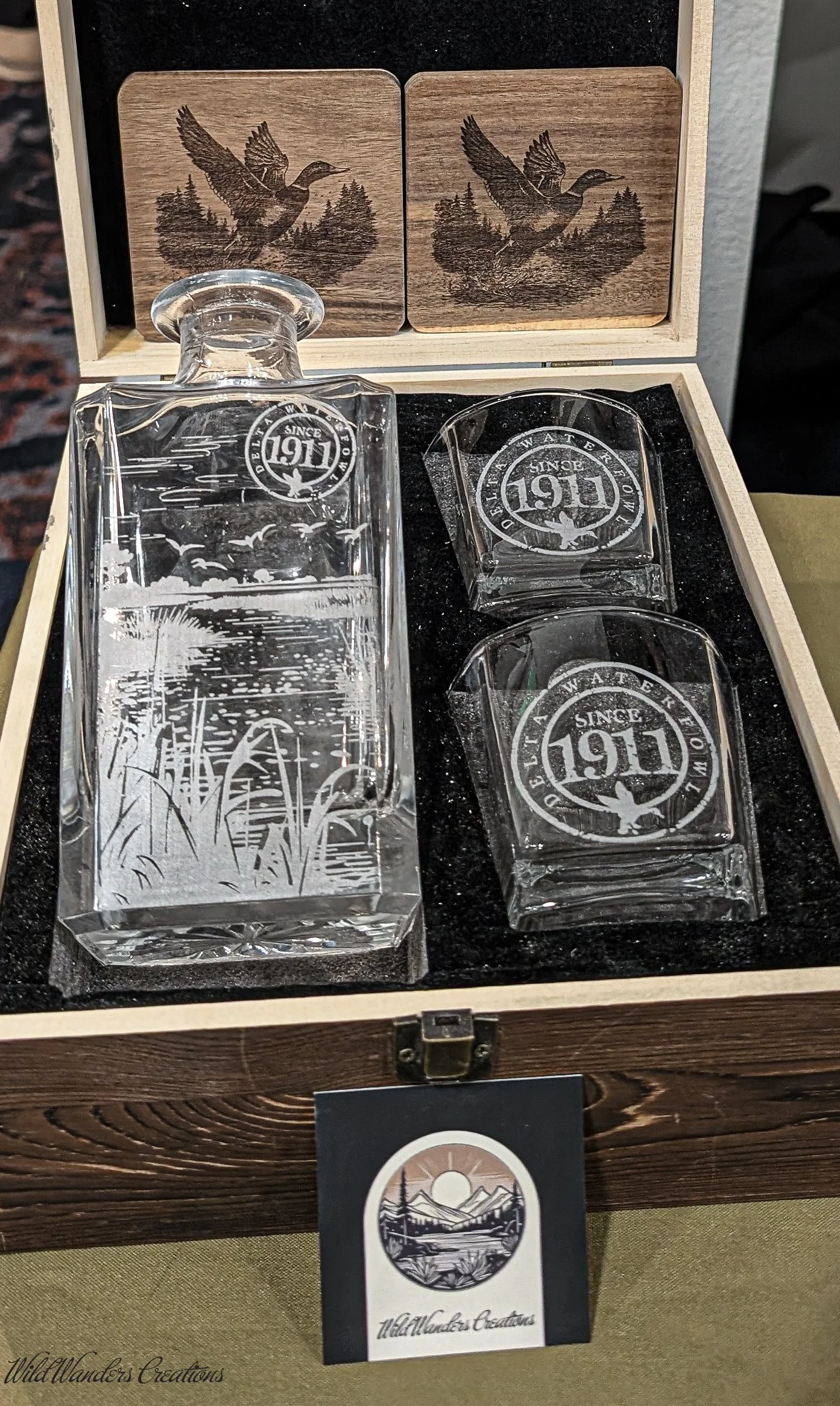Set of four glassware pieces and a wooden box with engraved images of a duck flying over water and nature scenes. The box contains a decanter with etched landscape and three matching cups with "Since 1911" and a duck logo.