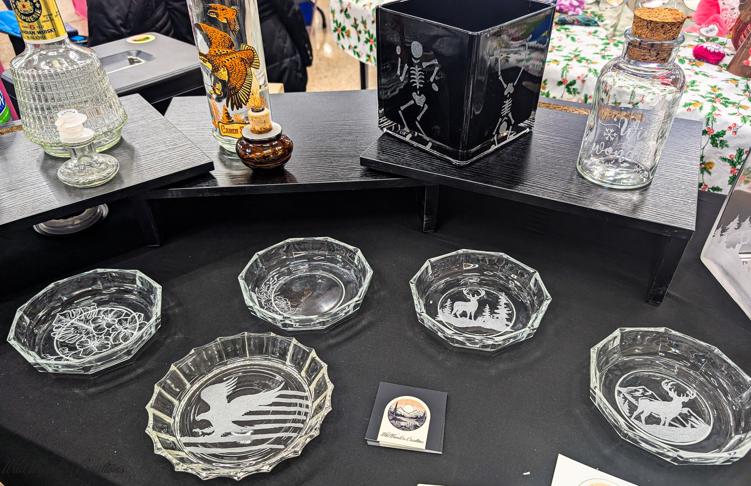 Decorative glass bowls with winter and nature motifs, displayed on a black tablecloth at an indoor craft market.