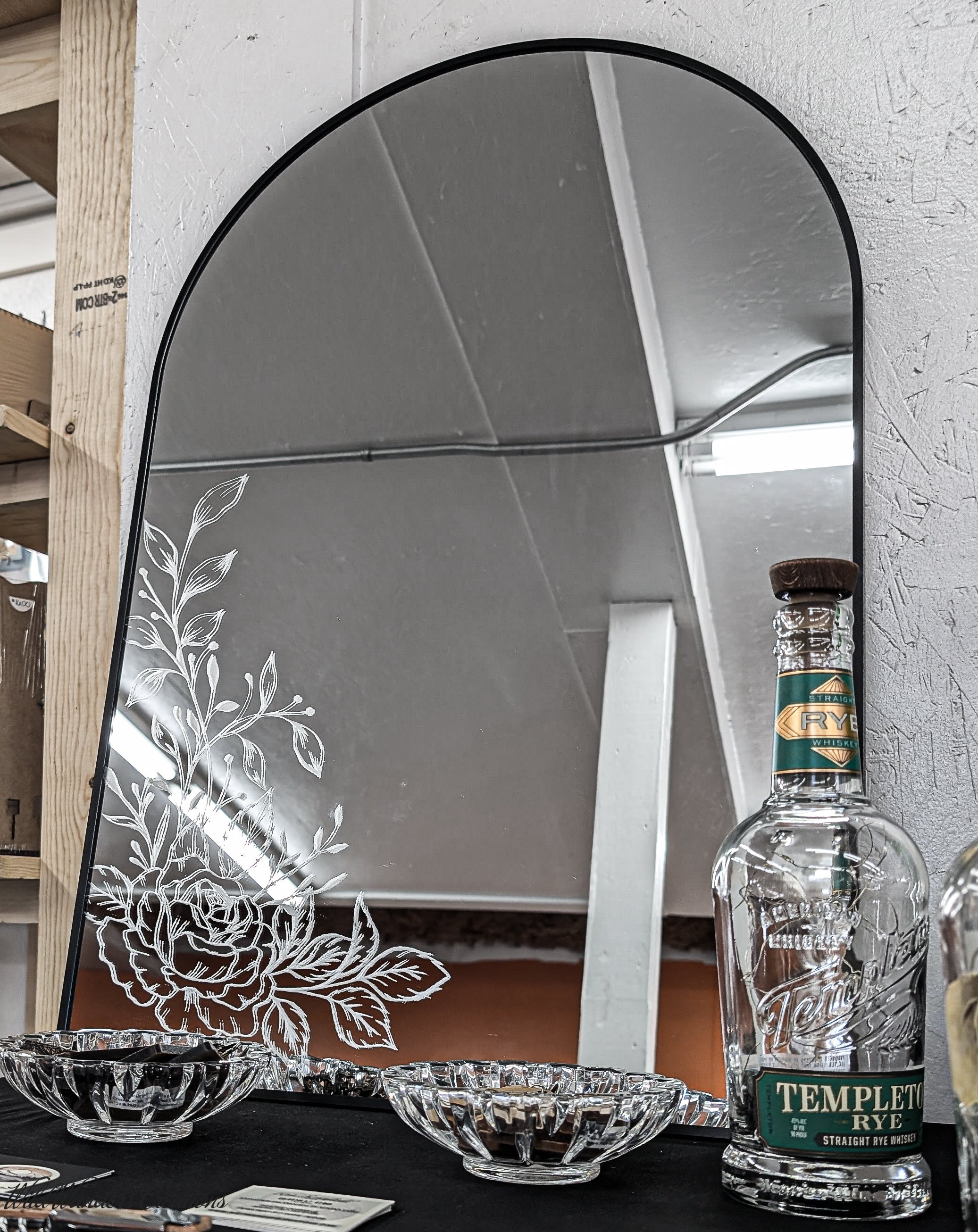 A decorative mirror with floral etching, on a table with glass bowls and a bottle of Templeton Rye whiskey, with a room reflected in the mirror.