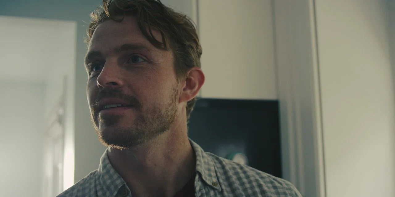 A man with light brown hair and a beard looks to the side in a room with natural light.