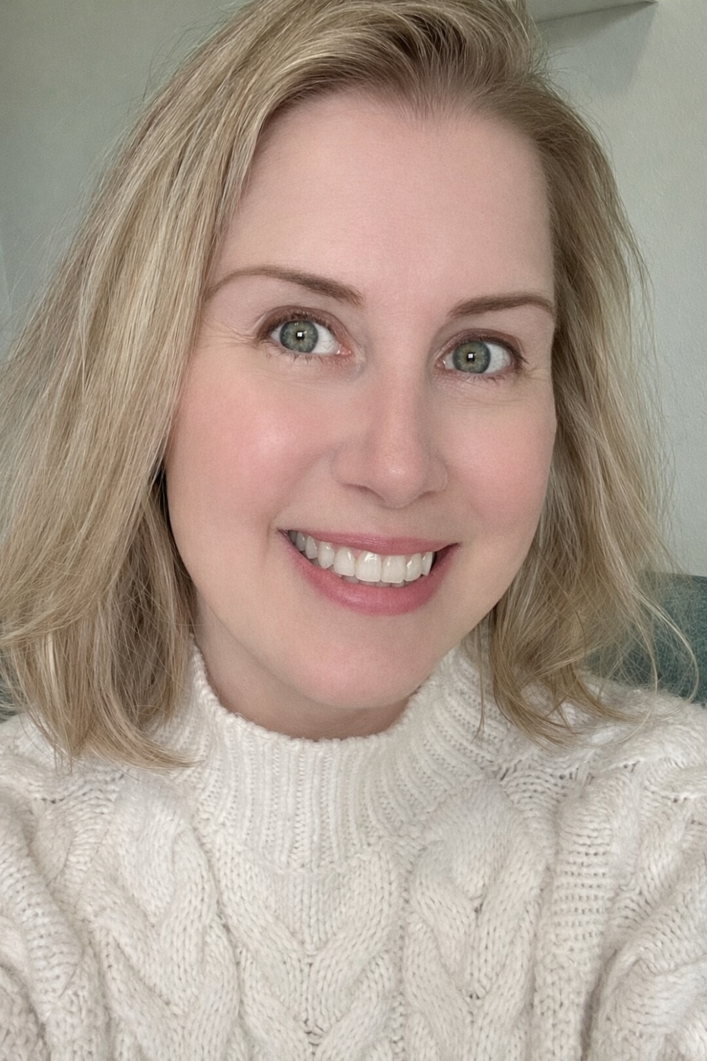 A smiling woman with blonde hair and blue eyes wearing a white knitted sweater.