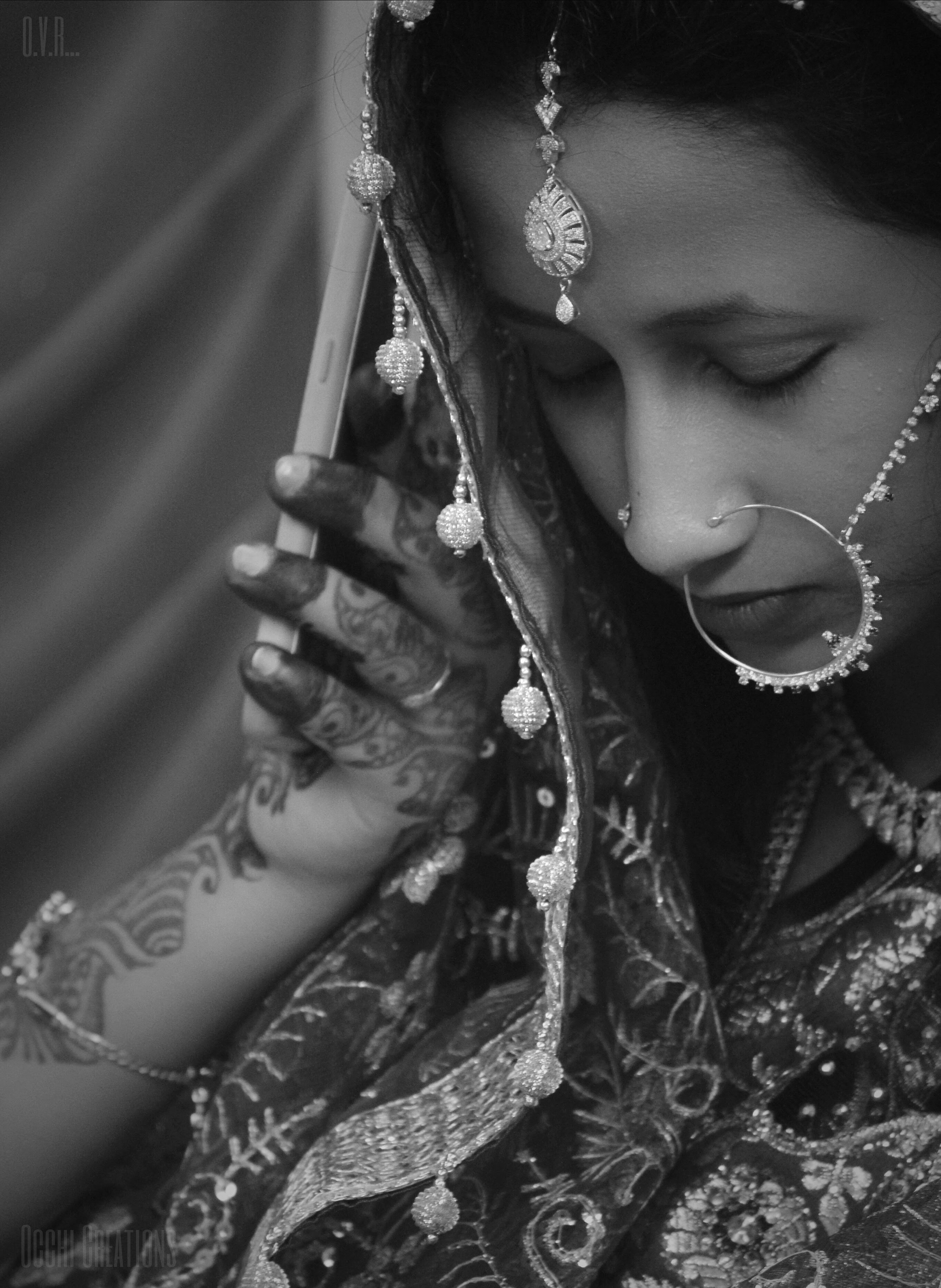 A woman dressed in traditional Indian attire with intricate jewelry, including a headpiece, nose ring, earrings, and bangles, holding a mobile phone close to her ear. She has henna designs on her hand and is looking down. The image is in black and wh