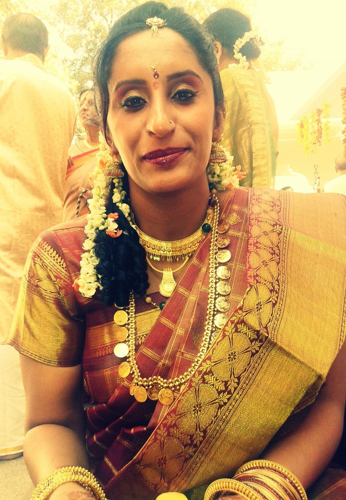 A woman dressed in traditional Indian attire at a celebration or wedding, adorned with gold jewelry, flowers, and a sari, smiling at the camera.