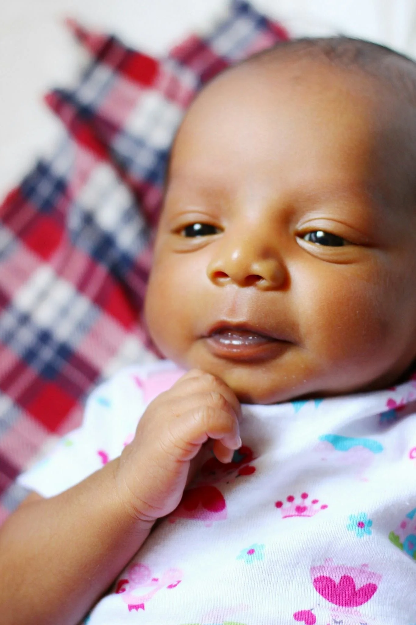 Close-up of a baby with dark hair and a cheerful expression, wearing a white outfit with colorful patterns, resting on a red and blue checkered blanket.