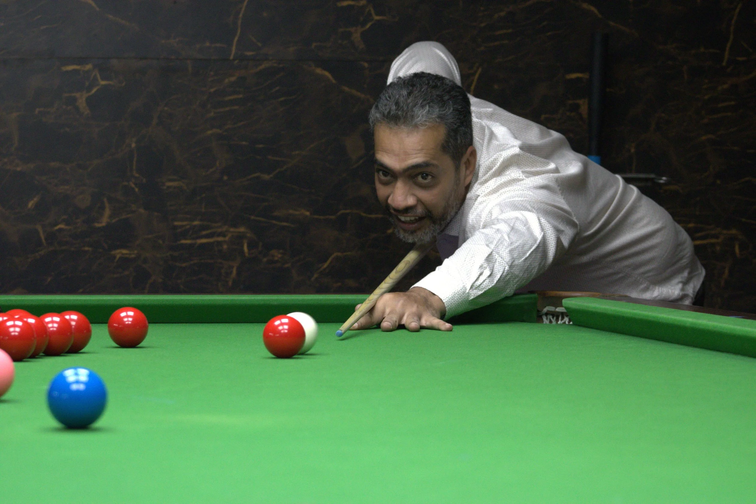 A man playing billiards, leaning over a green pool table with red, blue, and pink balls, aiming with a cue stick.