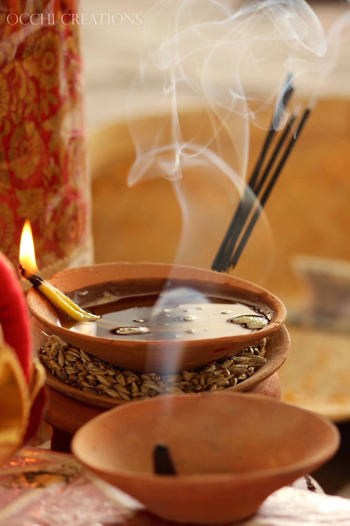 A close-up of an incense stick burning in a clay dish, with incense smoke rising upward. The dish is placed on top of another dish filled with seeds, and there are other incense sticks in the background. The scene appears to be a religious or spiritu