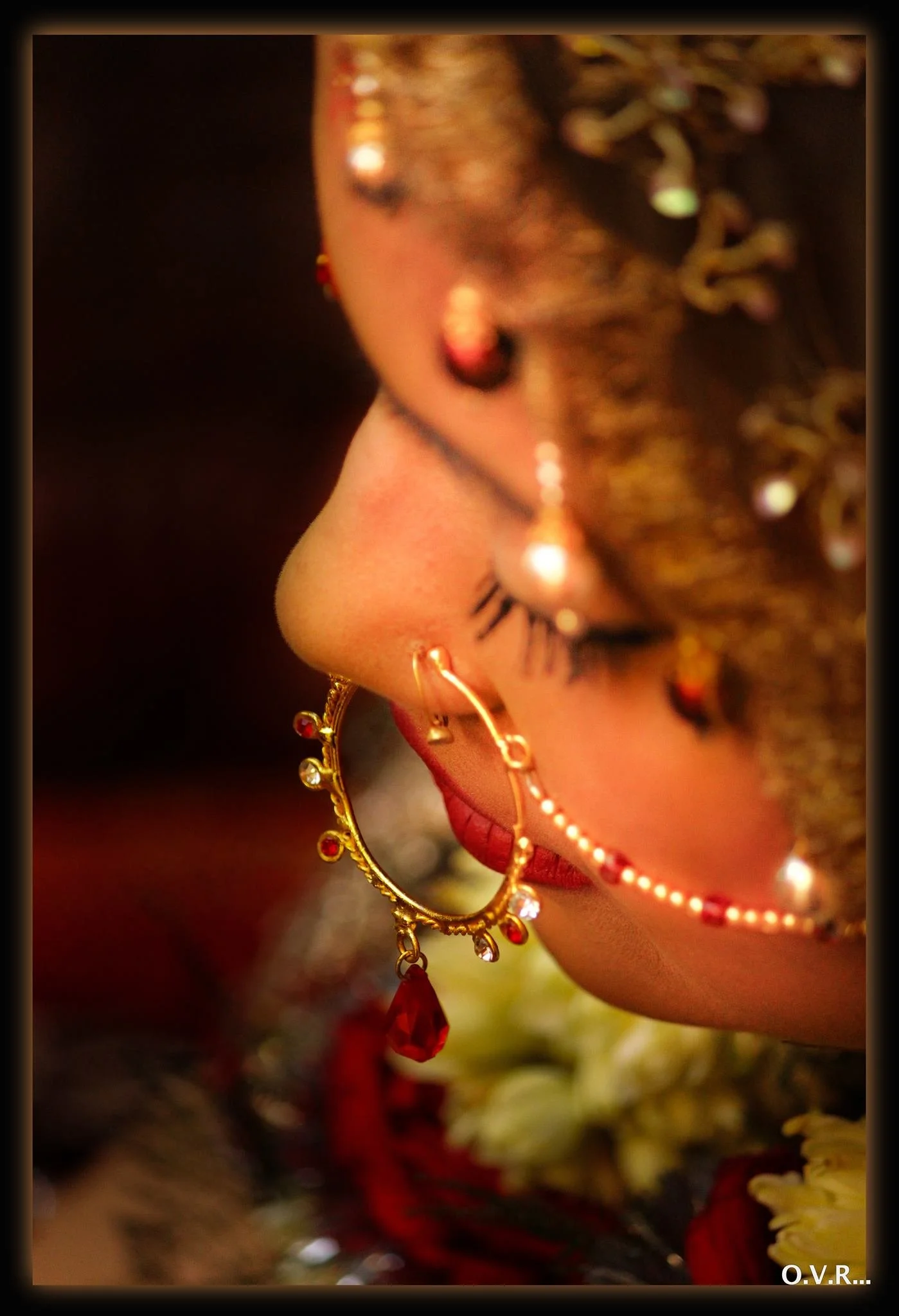 Close-up of a woman's face showing red lipstick and jewelry, including gold earrings with red gemstones and a decorative gold headpiece with beads, feathers, and gemstones.