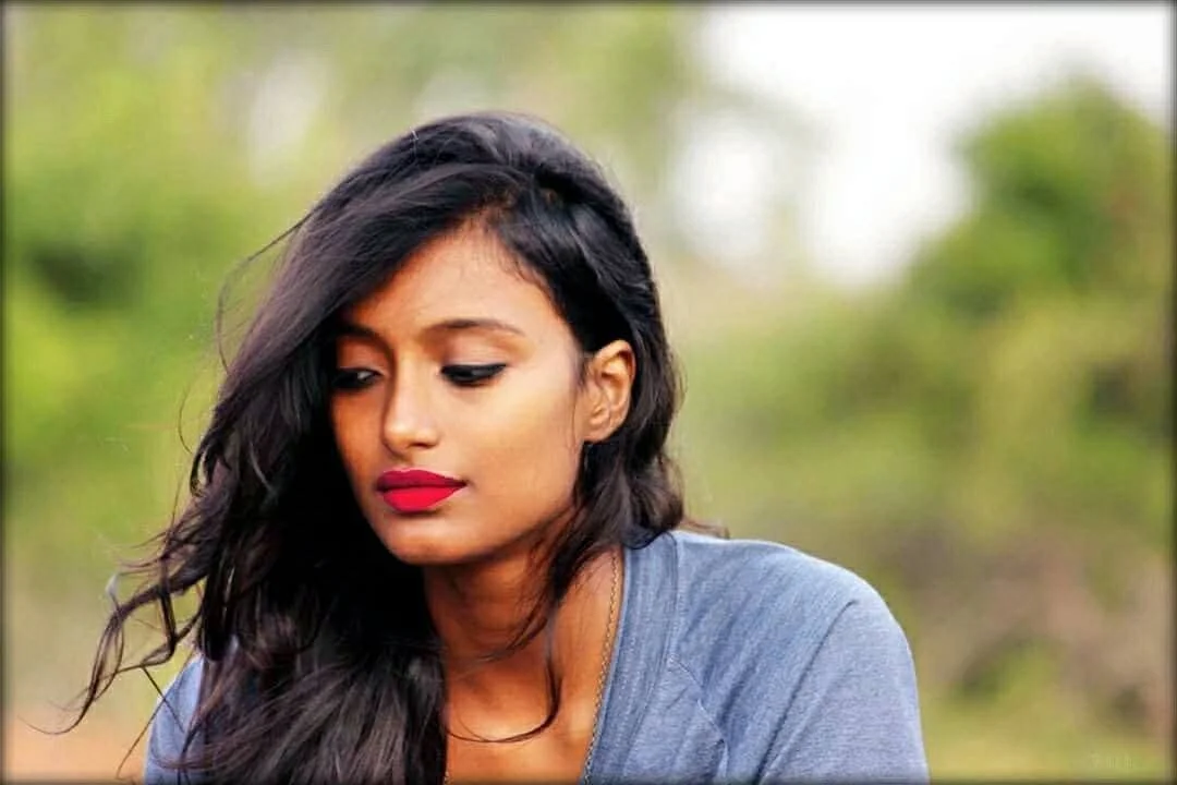 A young woman with long black hair, wearing a gray top and bright red lipstick, looking downward with a pensive expression, outdoors with a blurred green nature background.