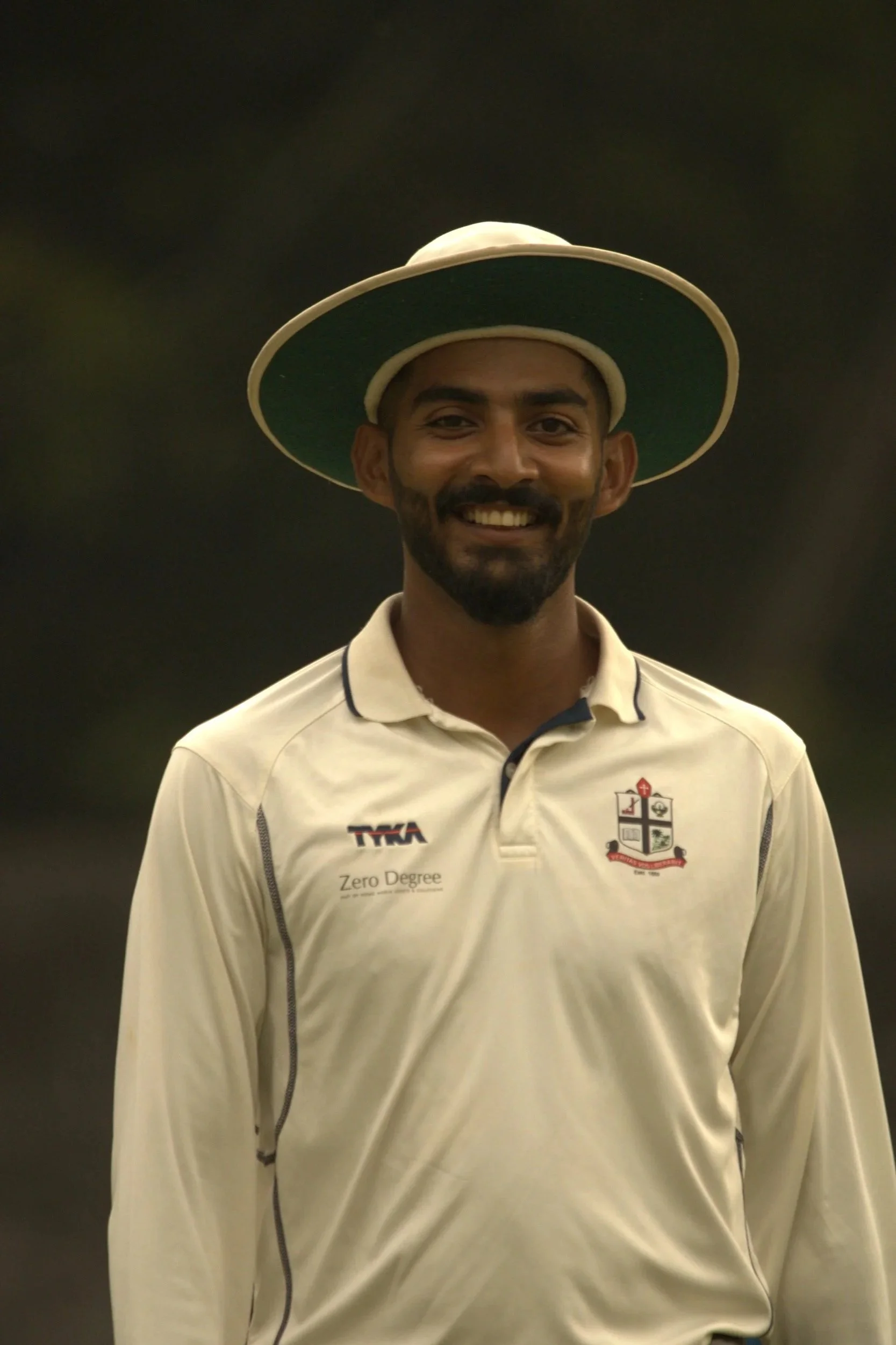 A man with a beard and mustache smiling, wearing a wide-brimmed green and white hat and a cream cricket jersey with various logos.