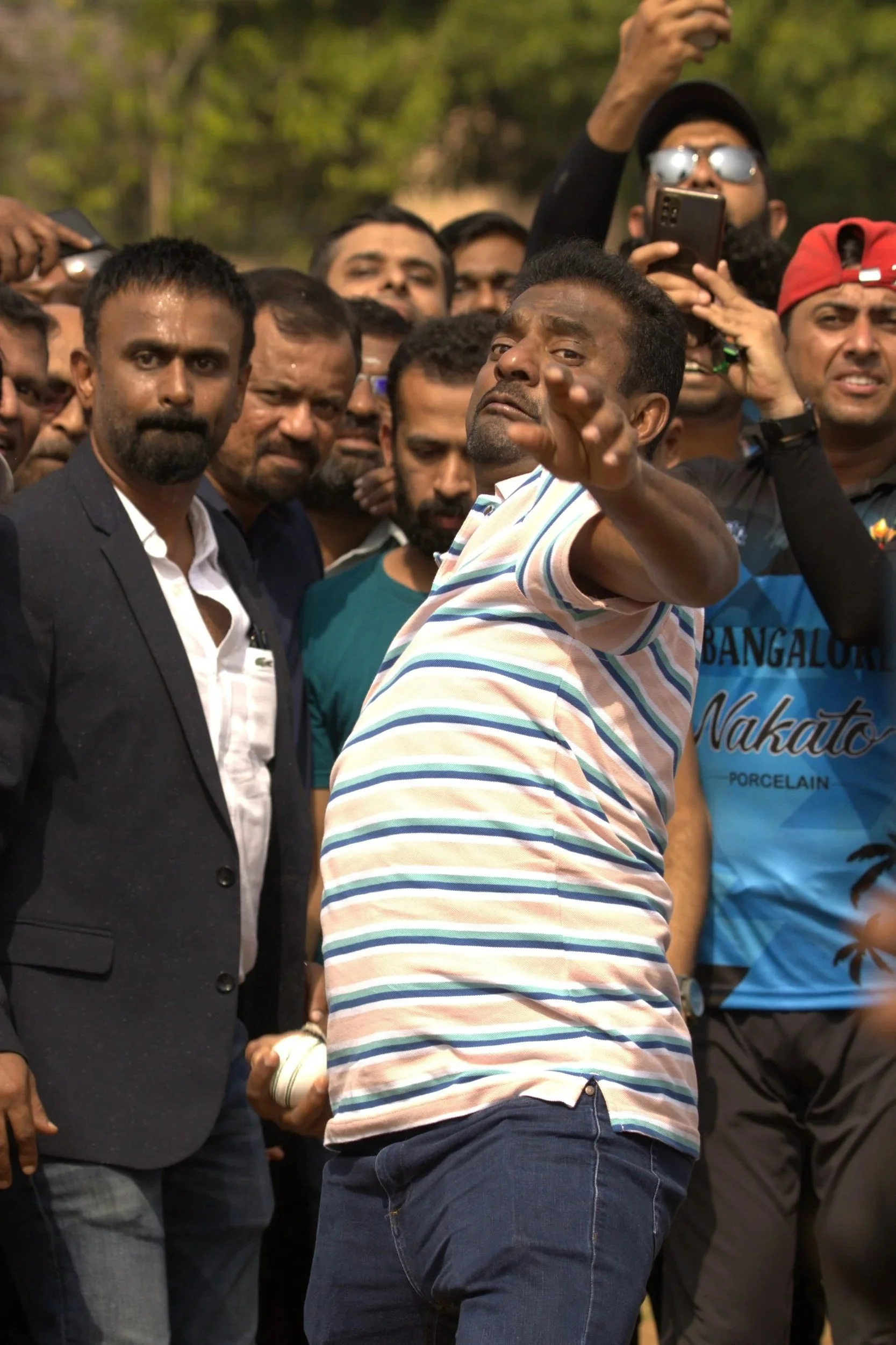 A man in a striped shirt pointing towards the camera surrounded by a group of people taking photos and watching him, outdoors with trees in the background.
