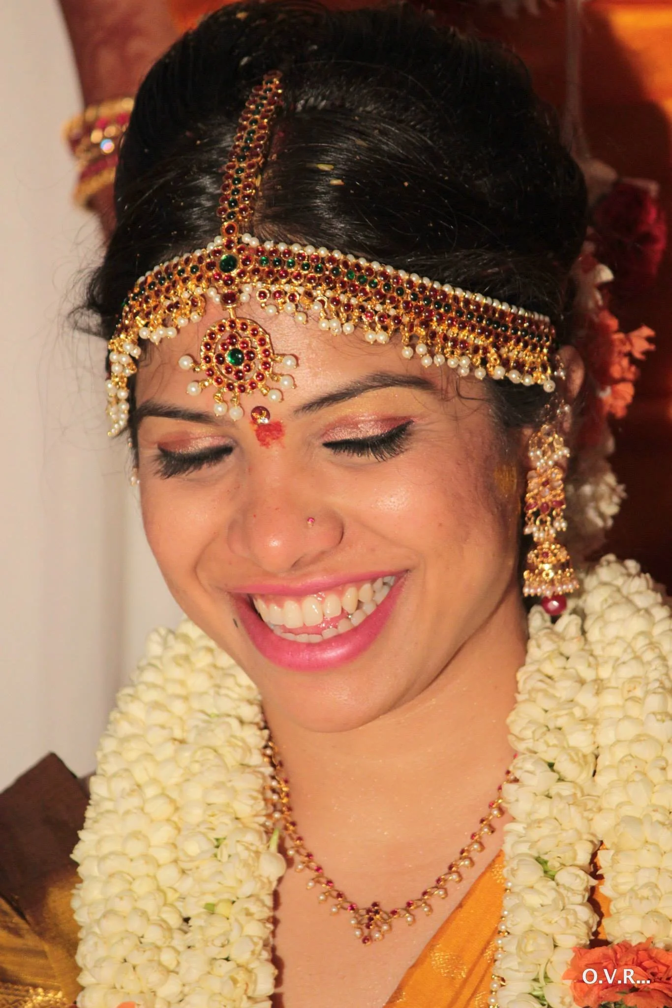 A woman dressed in traditional Indian wedding attire, smiling with closed eyes, wearing a gold and red saree, adorned with intricate jewelry and floral garlands.