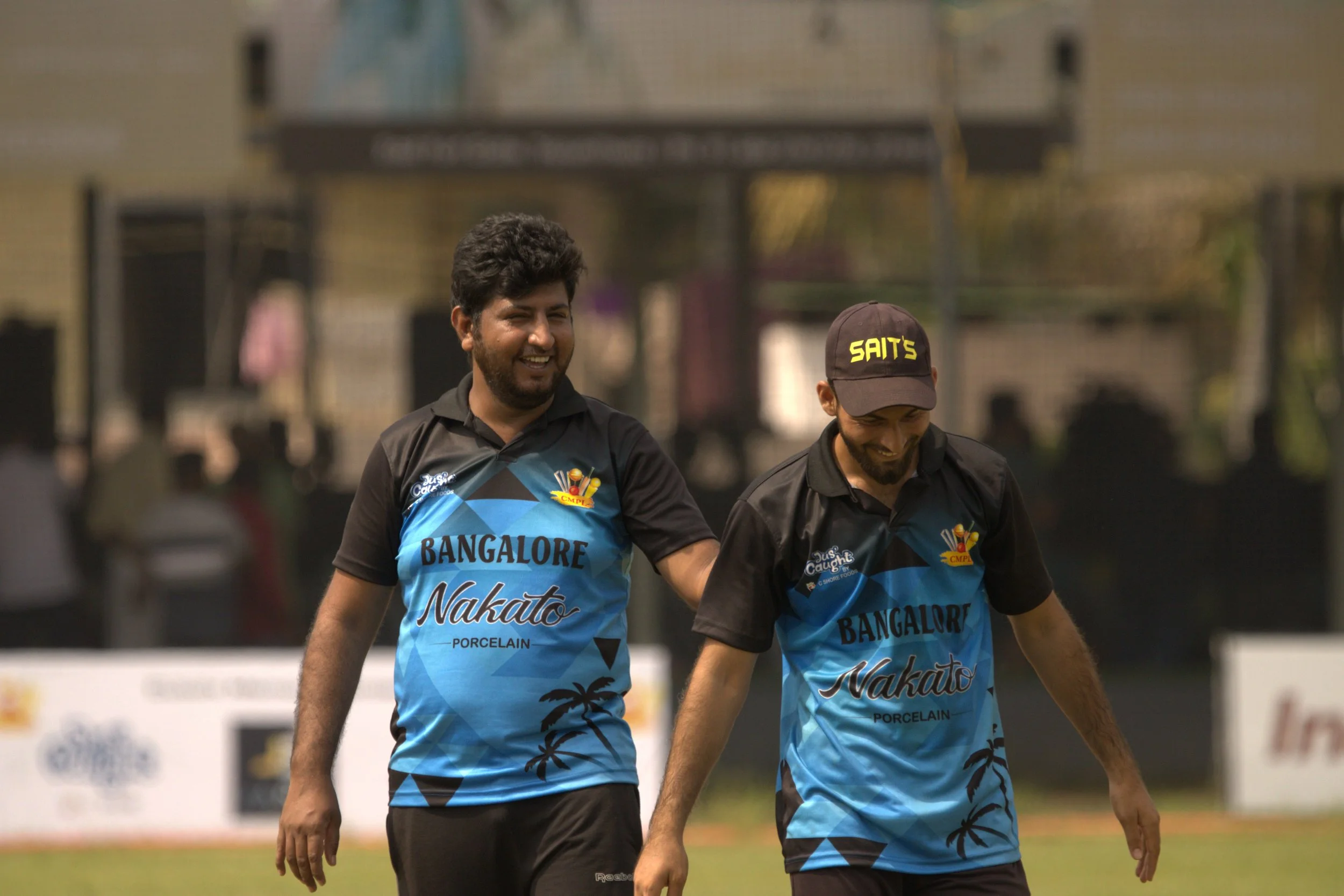 Two cricket players from Bangalore Nakato team walking together on the field, smiling and enjoying the game, wearing blue and black uniforms with the team name and sponsor logos.