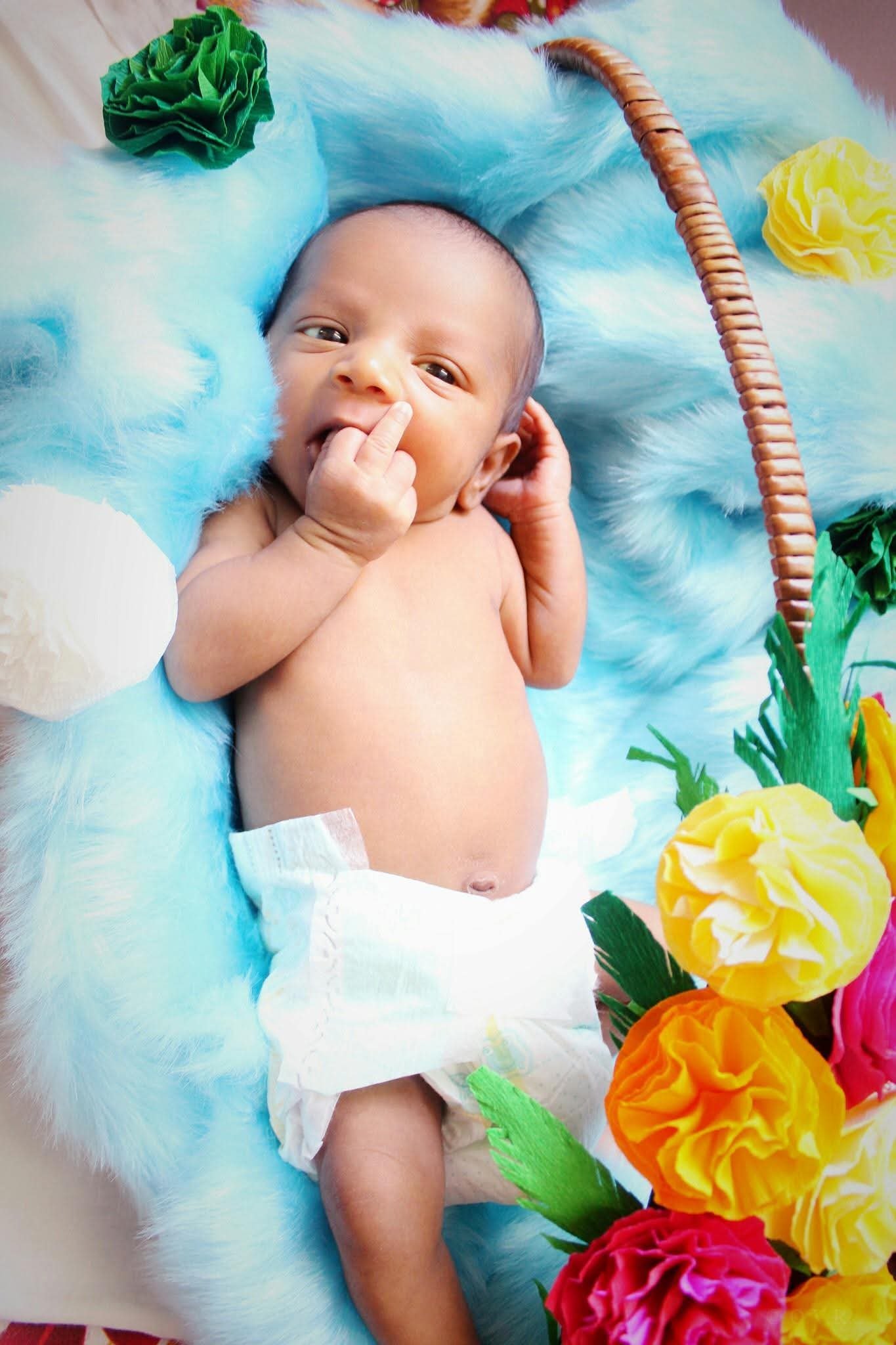 A baby lying on a fluffy blue blanket surrounded by colorful paper flowers, wearing a diaper, with one hand near their mouth and the other behind their head.