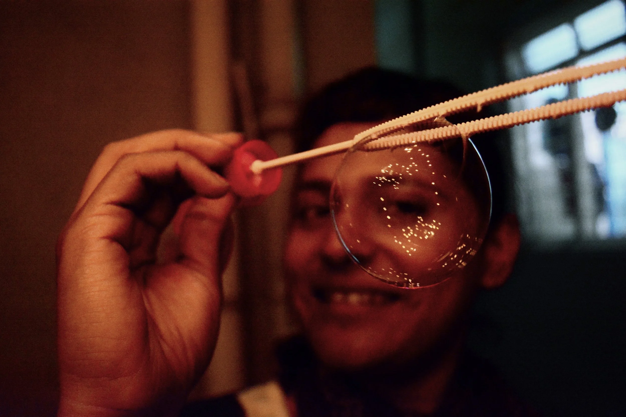 Person holding a bubble wand near their eye, with reflections of lights on the bubble, in a dimly lit indoor setting.