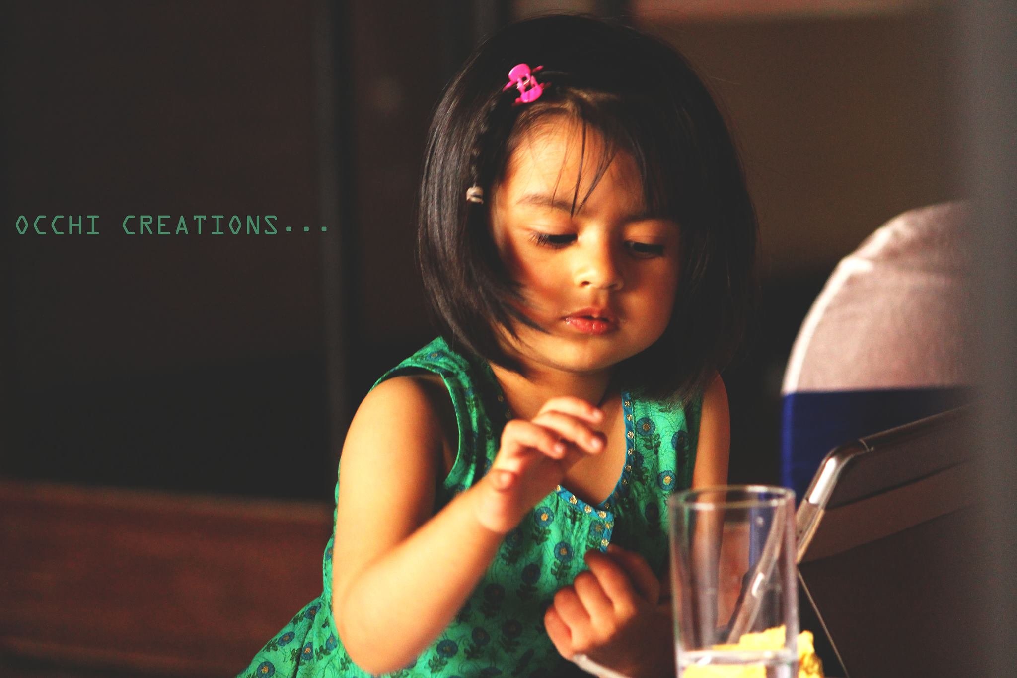 A young girl with short black hair and a pink hair clip, wearing a green dress, sitting at a table in front of a tablet and a glass of water, looking at the screen with a focused expression.