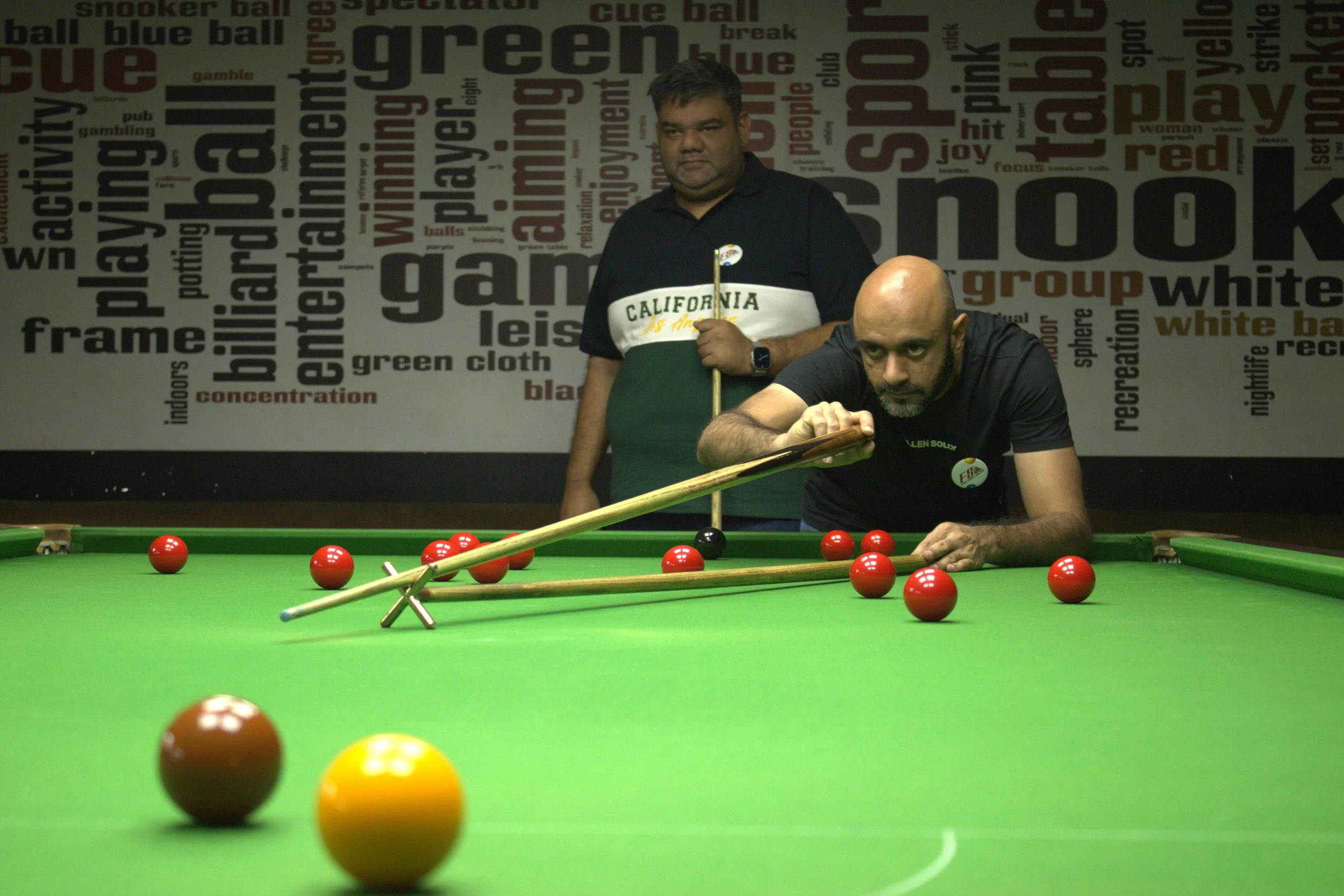Two men playing carom billiards on a green table, one striking the cue ball with a cue stick while the other stands behind watching, with a background featuring words related to billiards and snooker.