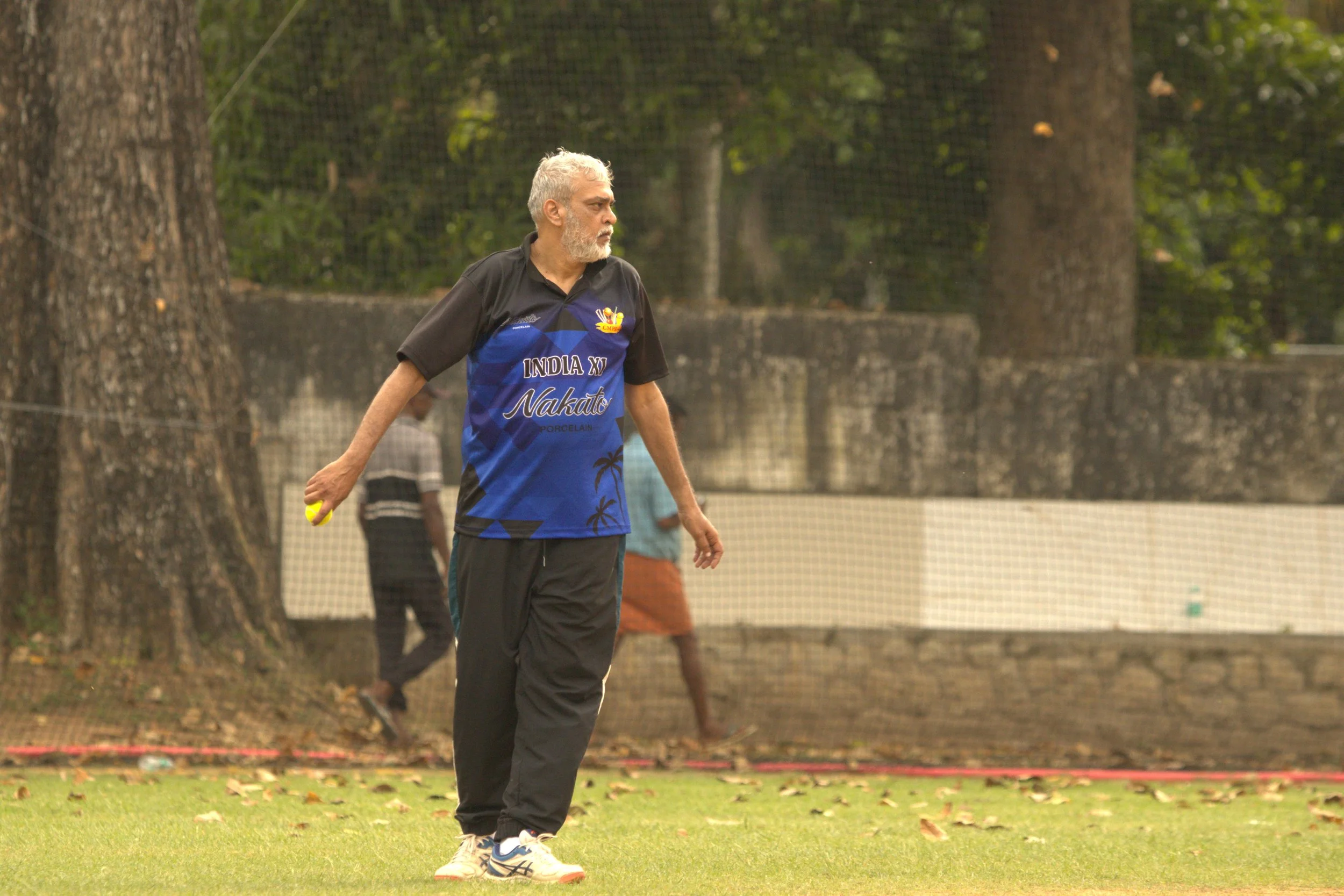 An older man with white hair and beard holding a tennis ball standing on a grass field. He is wearing a dark and blue sports shirt with the text 'India XI Nakato' and black pants. There are large trees and a net in the background.