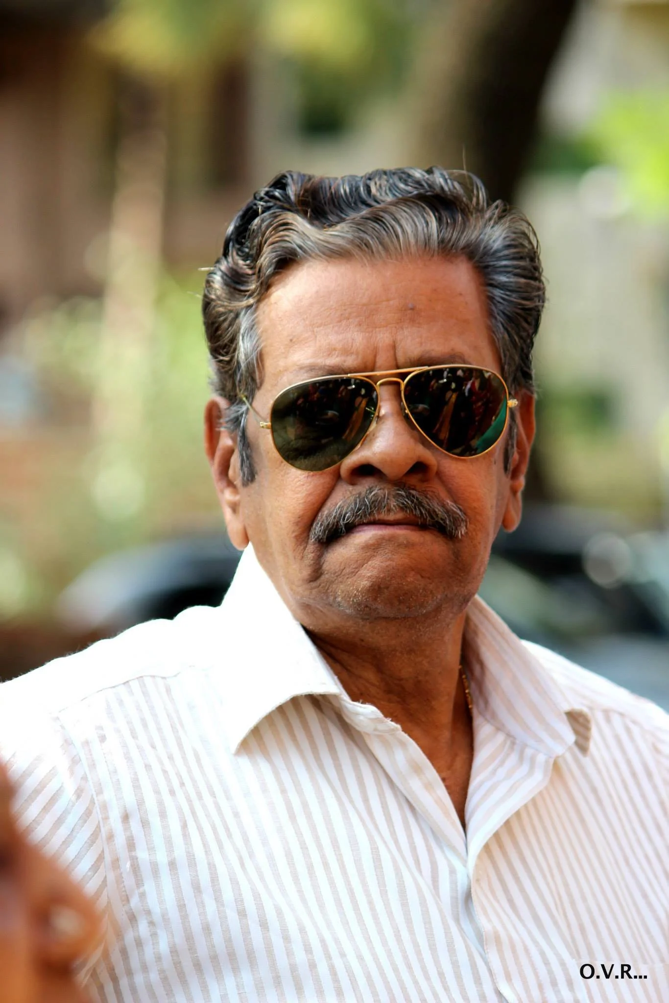 A man with salt-and-pepper hair, mustache, and tan skin wearing a white shirt and dark sunglasses stands outdoors with a blurred background of trees and sunlight.