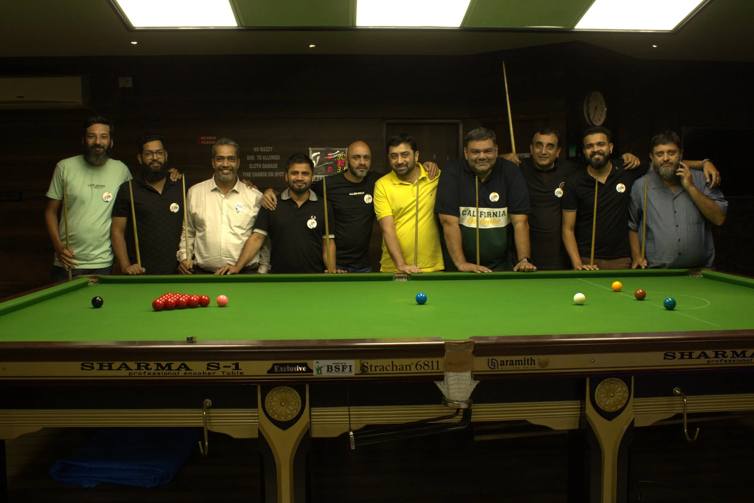 Group of eleven men standing behind a snooker table in an indoor setting, some with their arms around each other, smiling, with colored balls and cues on the table.