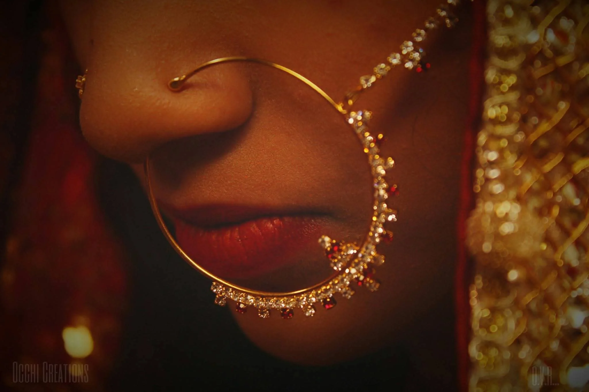 Close-up of a woman's face with a gold hoop earring adorned with red and clear gemstones, earrings, and gold jewelry, with focus on her nose and lips, showing her skin tone and jewelry details.