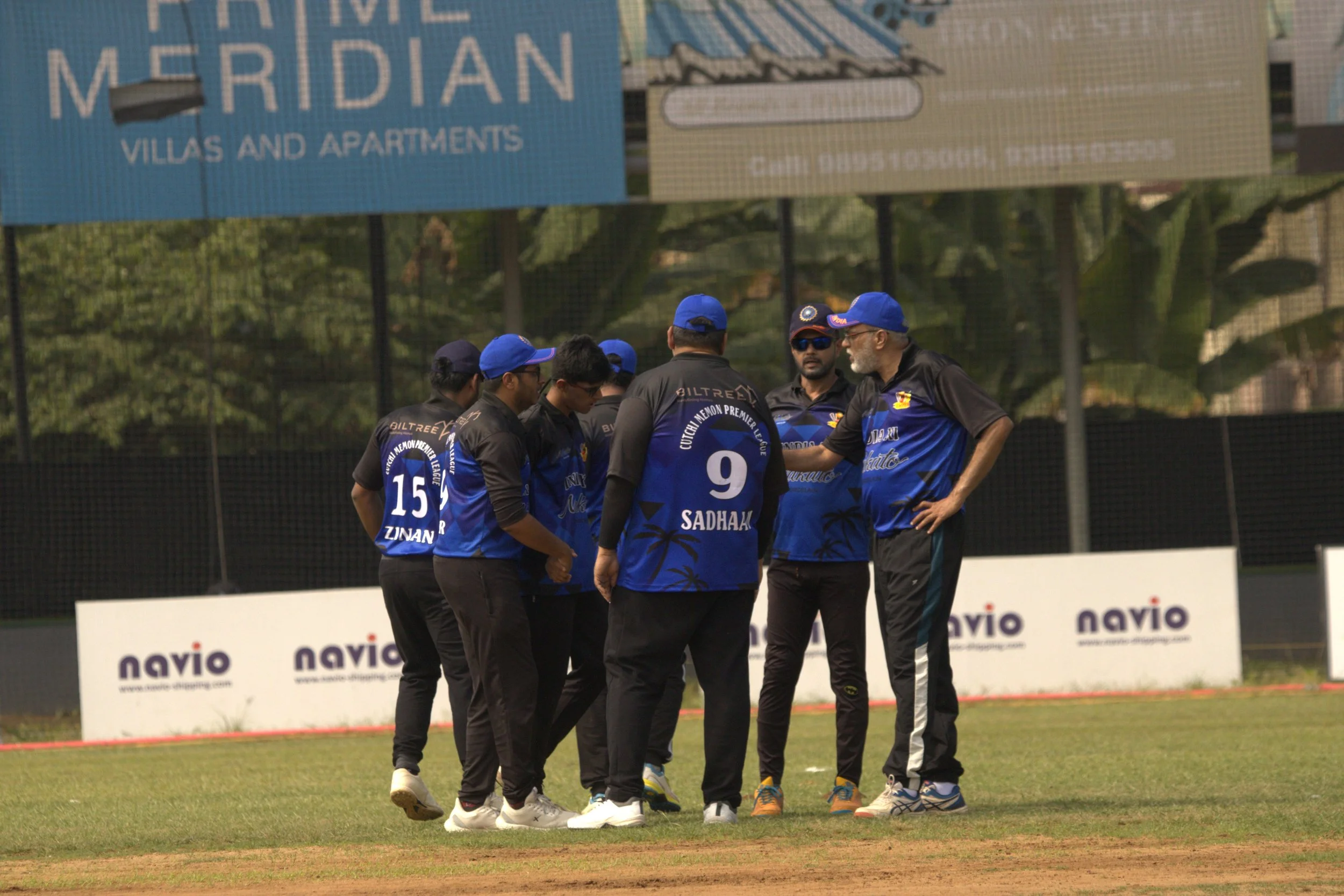 A group of cricket players dressed in black and blue uniforms is gathered on a cricket field during a game. They are engaged in a discussion, with some wearing caps and sunglasses.