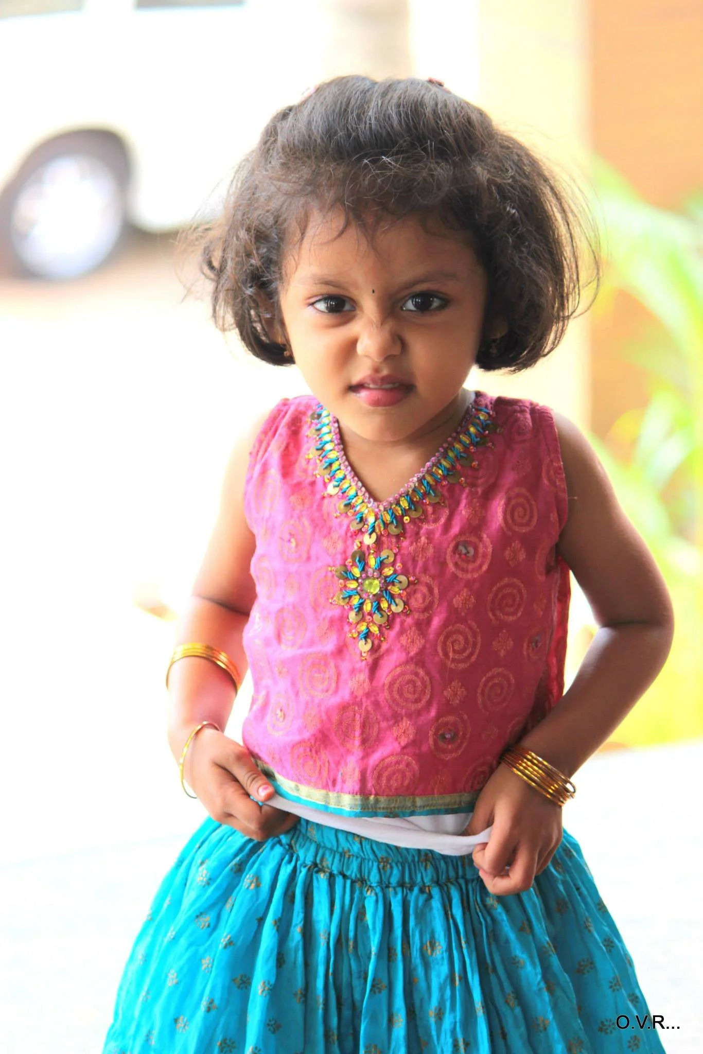A young girl with dark, curly hair and an angry or confused facial expression, wearing a pink and multicolored traditional Indian dress and gold jewelry.