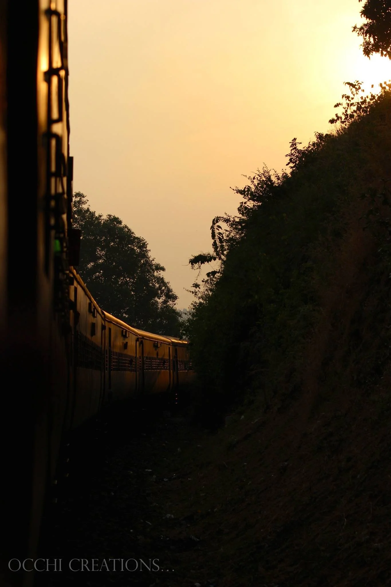 A train is moving along a curved track beside a hillside, with trees and a sunset sky in the background.