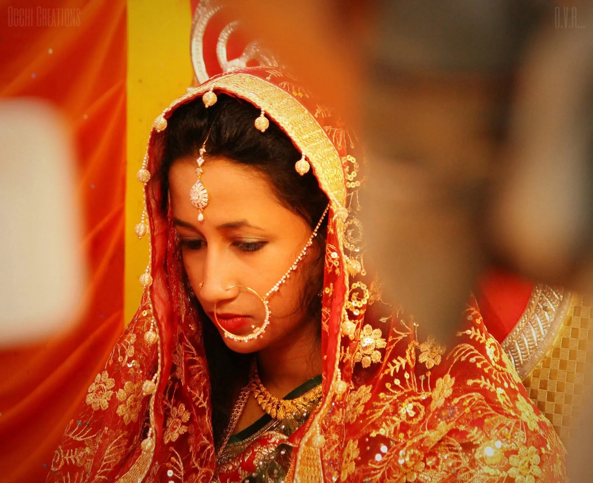 A woman dressed in traditional Indian bridal attire, wearing a red and gold embroidered saree, with jewelry including a maang tikka, nose ring, earrings, and necklace, standing indoors.