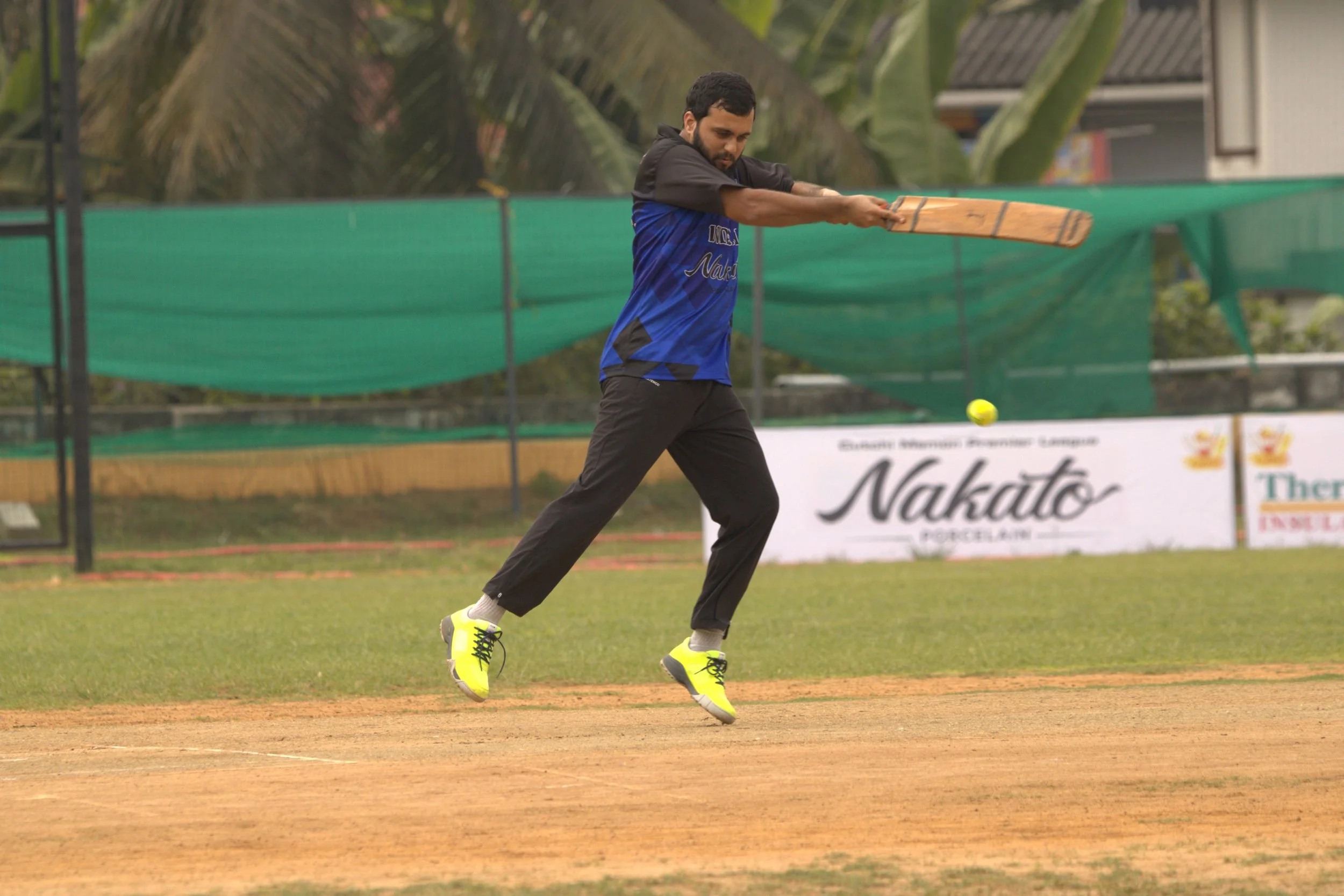 A man playing cricket on a field, wearing yellow sneakers, blue and black jersey, holding a cricket bat, mid-action, with a background of green fencing and trees.