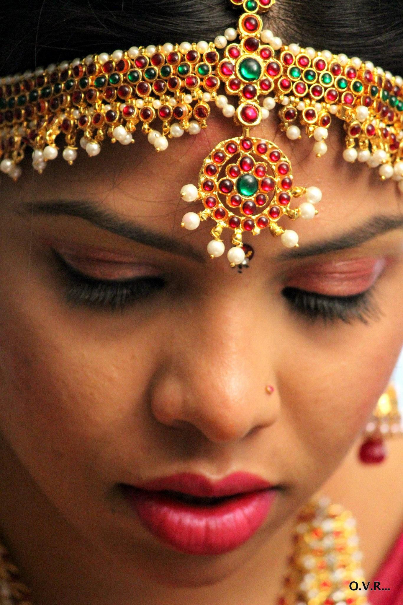 Close-up of a woman wearing traditional Indian jewelry with intricate gold and red designs, pearls, and green accents, including a forehead ornament and earrings.