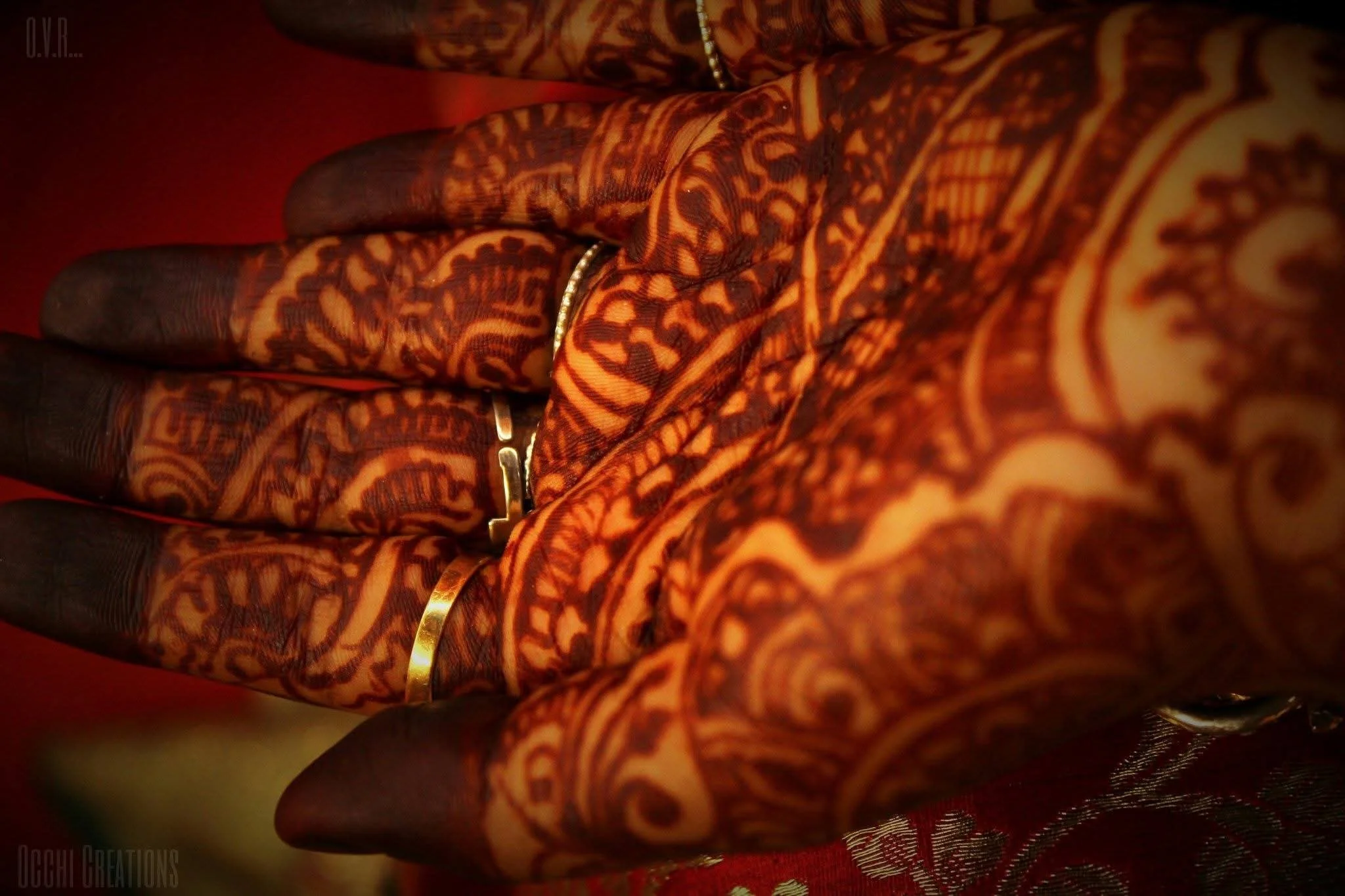 Close-up of a hand with intricate henna (mehndi) designs and wearing gold rings, set against a red background.