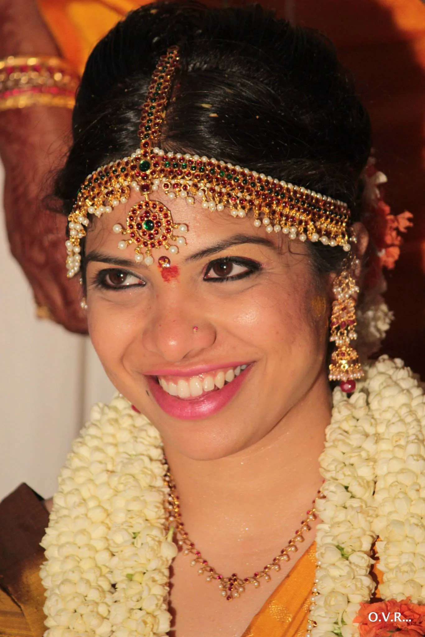 Close-up of a woman dressed in traditional Indian bridal attire, wearing ornate jewelry including a headpiece, earrings, necklace, and floral garlands, smiling joyfully.
