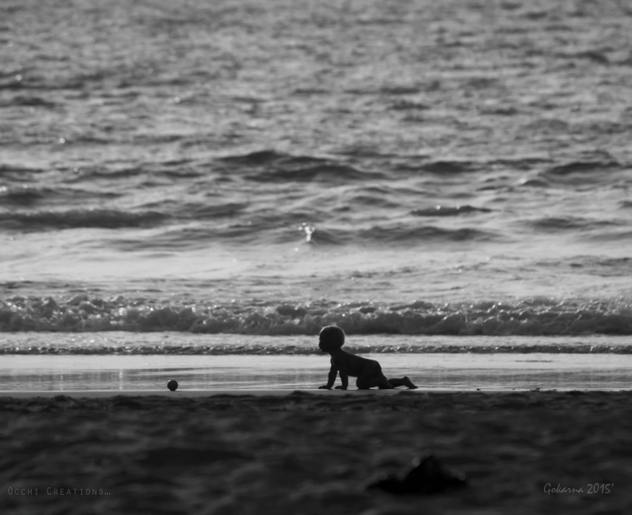 A black and white photo of a child crawling on the beach near the shoreline, with the ocean waves behind them and a small ball nearby.
