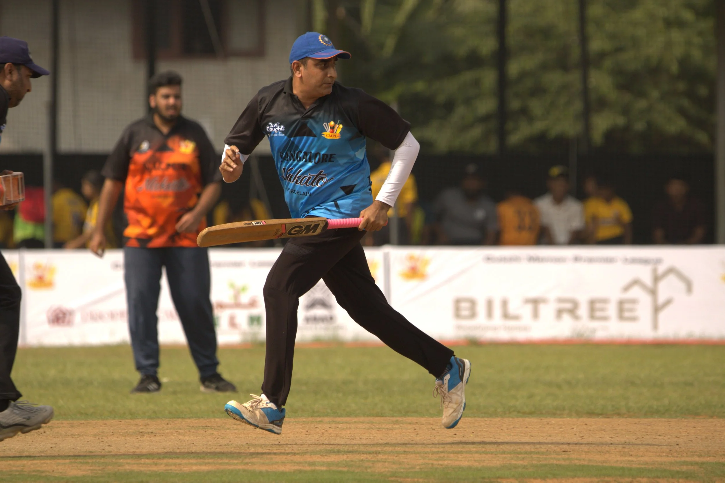 Cricket player in black and blue uniform running on the field holding a cricket bat, with other players and spectators in the background.