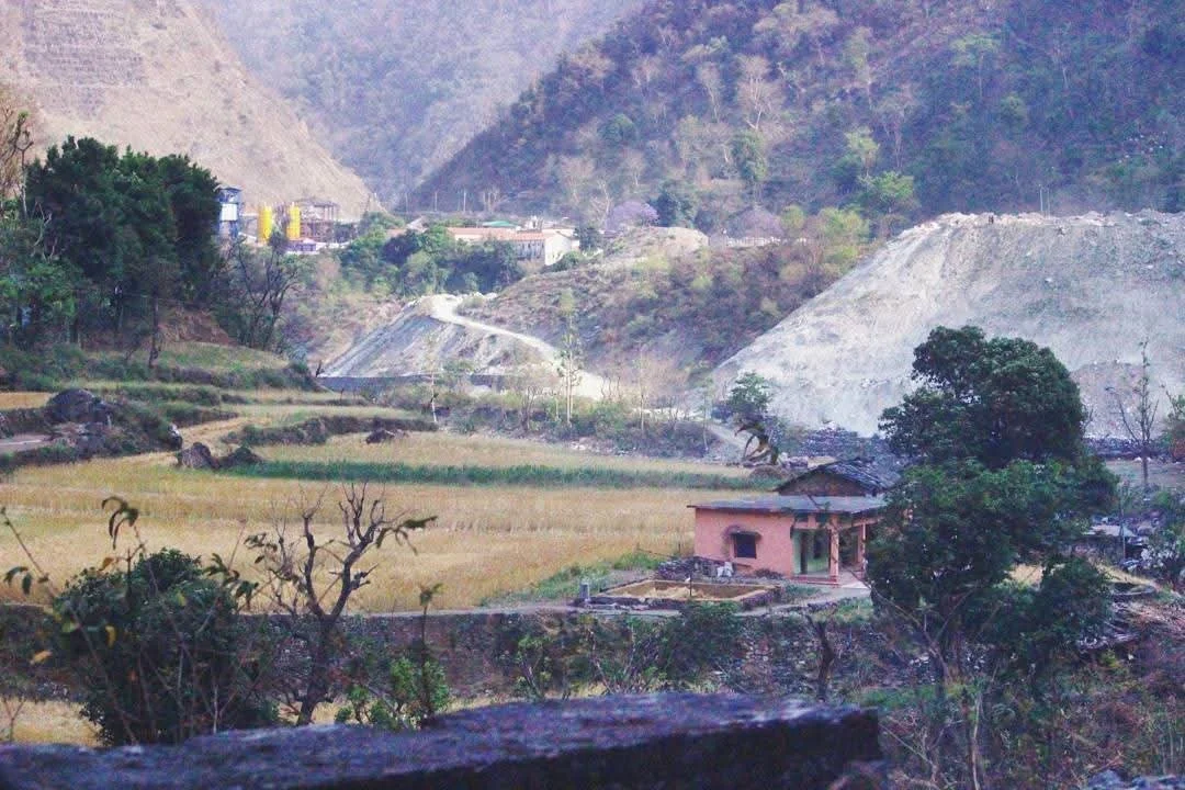 Scenic mountain landscape with terraced fields, sparse trees, a small pink house, and distant mountains with some construction or excavation activity.
