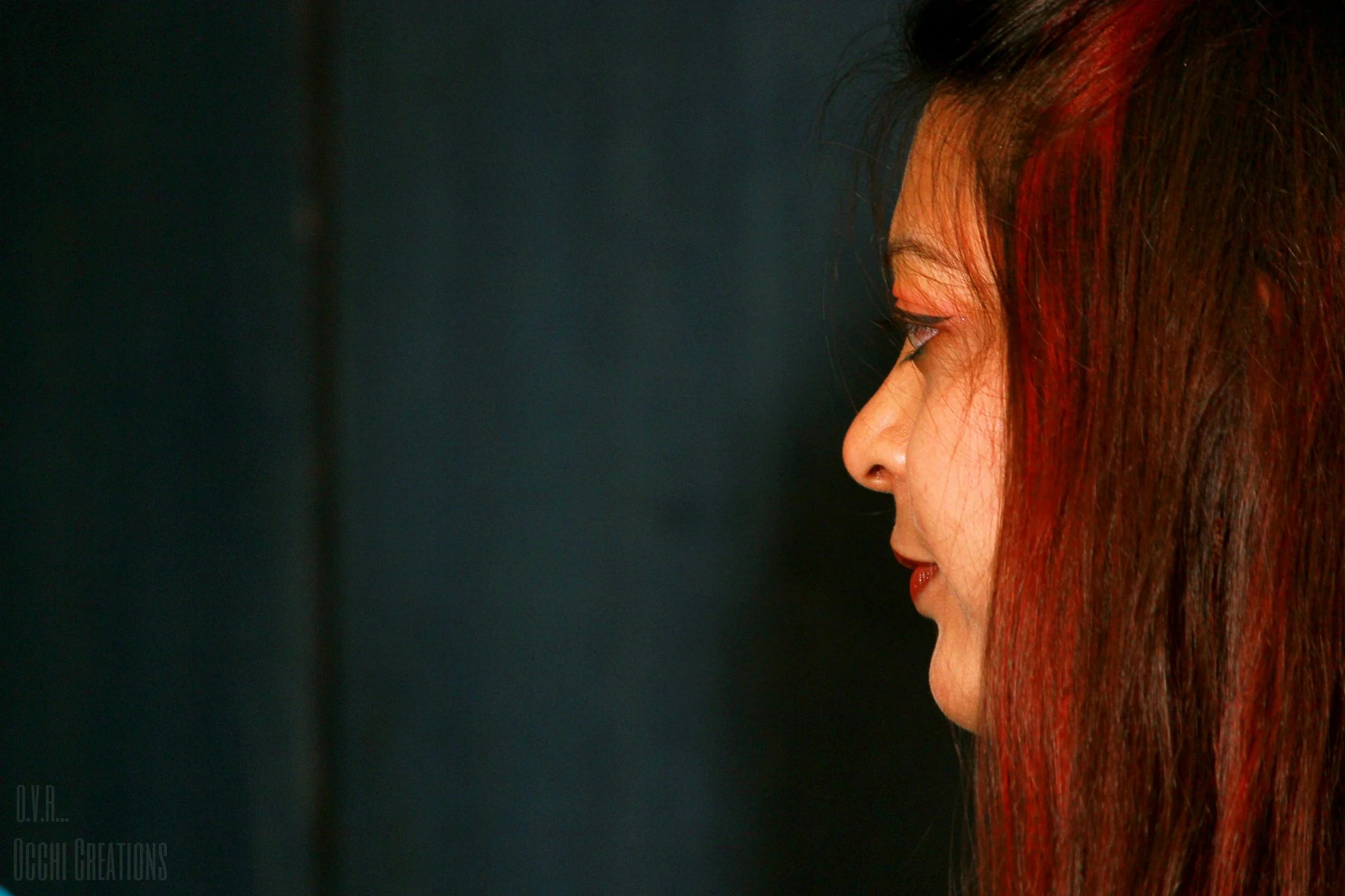 Profile of a woman with long, dark hair with red highlights, seen against a dark background.