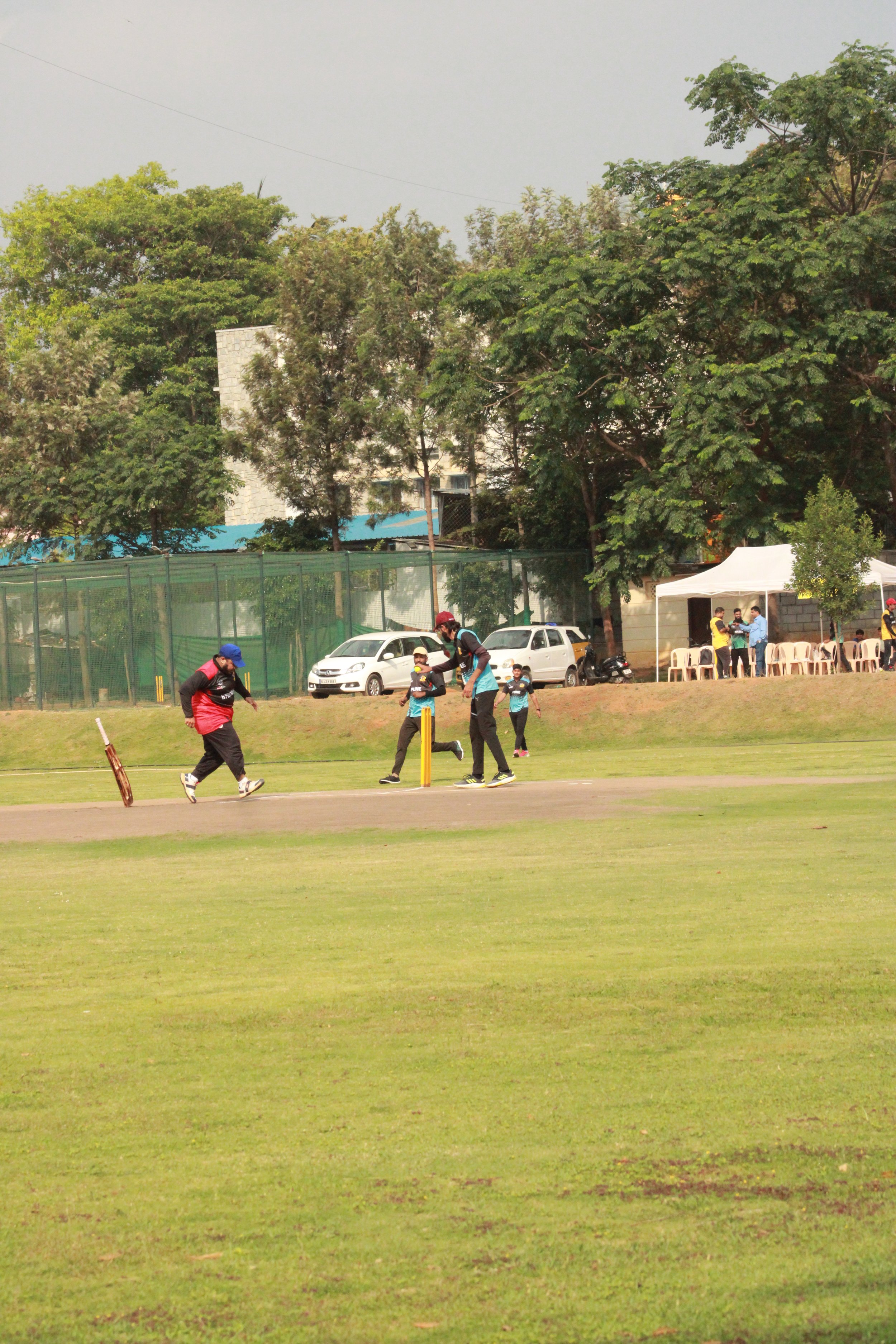 People playing cricket on a grassy field in a park with trees, a tent, and cars parked in the background.